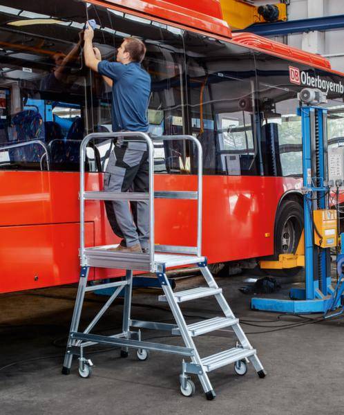 Ein Mann steht auf einer Trittleiter und führt Wartungsarbeiten an einem roten Bus durch. Im Hintergrund sind Werkstattmerkmale erkennbar.