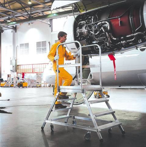 Ein Techniker in gelbem Overall arbeitet auf einer Leiter an einem geöffneten Flugzeugtriebwerk in einer großen, hellen Werkstatt.