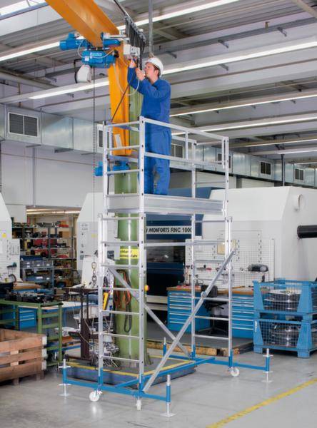 Ein Arbeiter in blauer Kleidung steht auf einem Gerüst in einer Industriehalle und bedient ein Gerät an einem hohen, orangefarbenen Pfeiler.