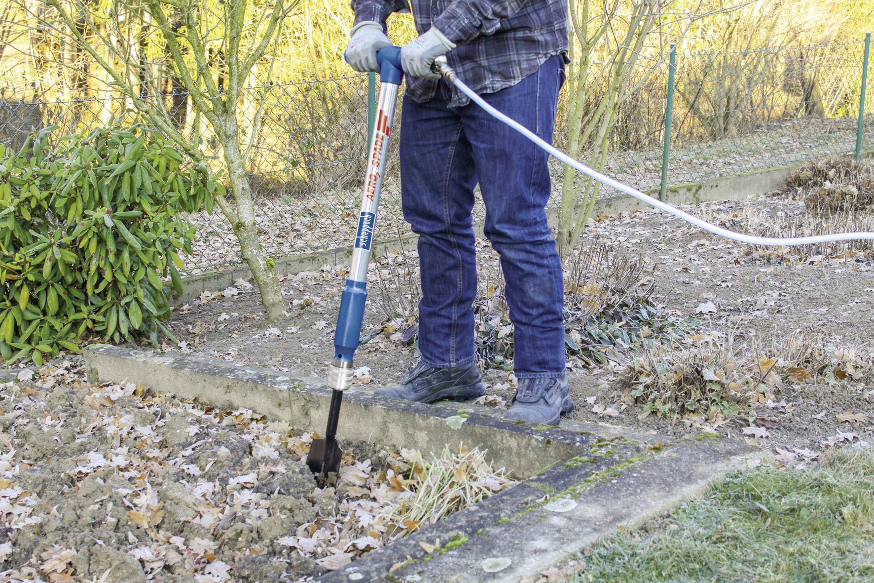 Eine Person belüftet den Boden eines Gartens mit einem Handgerät. Sie trägt Handschuhe und steht zwischen kahlen Beeten.