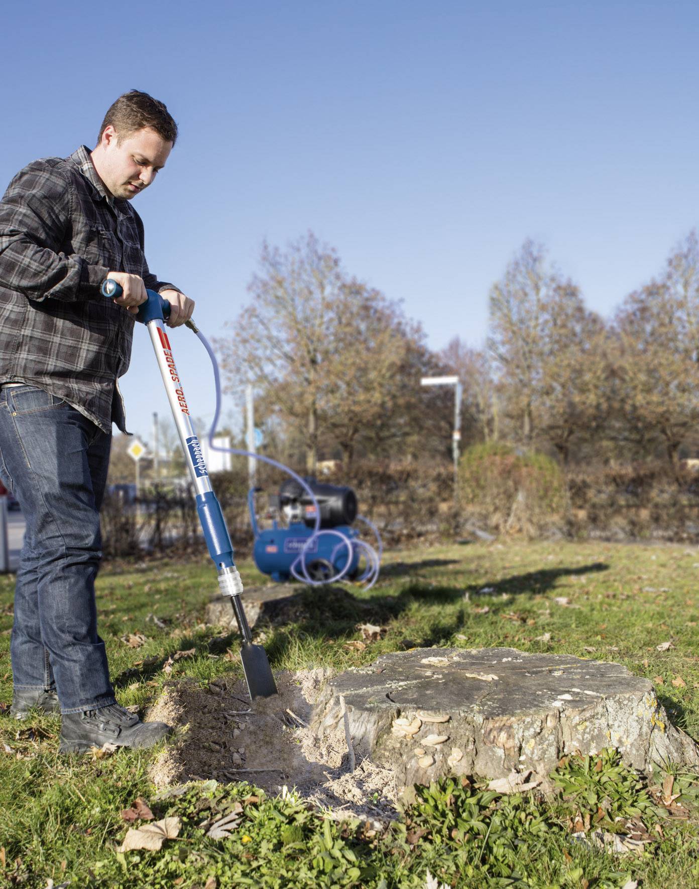 Ein Mann benutzt ein motorisiertes Werkzeug, um einen Baumstumpf in einem Garten zu entfernen. Im Hintergrund sind Bäume und ein Schlauch sichtbar.