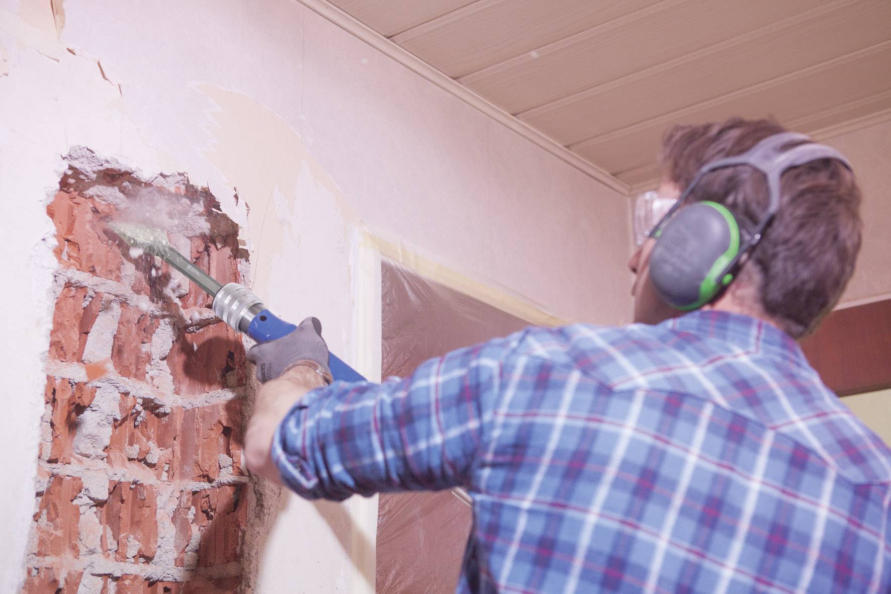 Mann in kariertem Hemd und Gehörschutz arbeitet mit einem Bohrhammer eine Wand auf. Teile der Wand zeigen offene Backsteine.