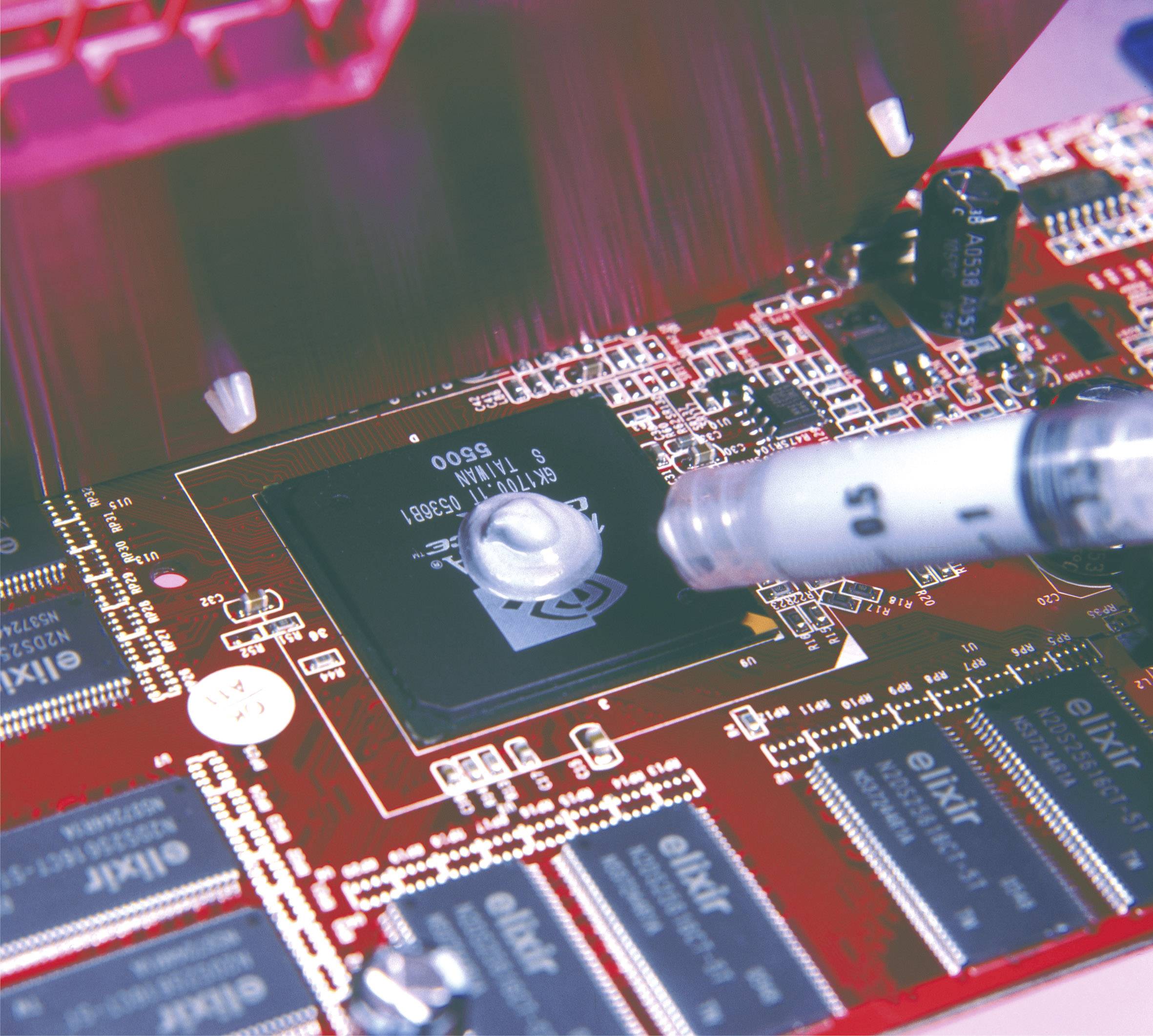 Nahaufnahme einer Computerplatine, auf der eine Paste auf einen Chip aufgetragen wird. Die Platine zeigt verschiedene elektronische Bauteile und Leiterbahnen.