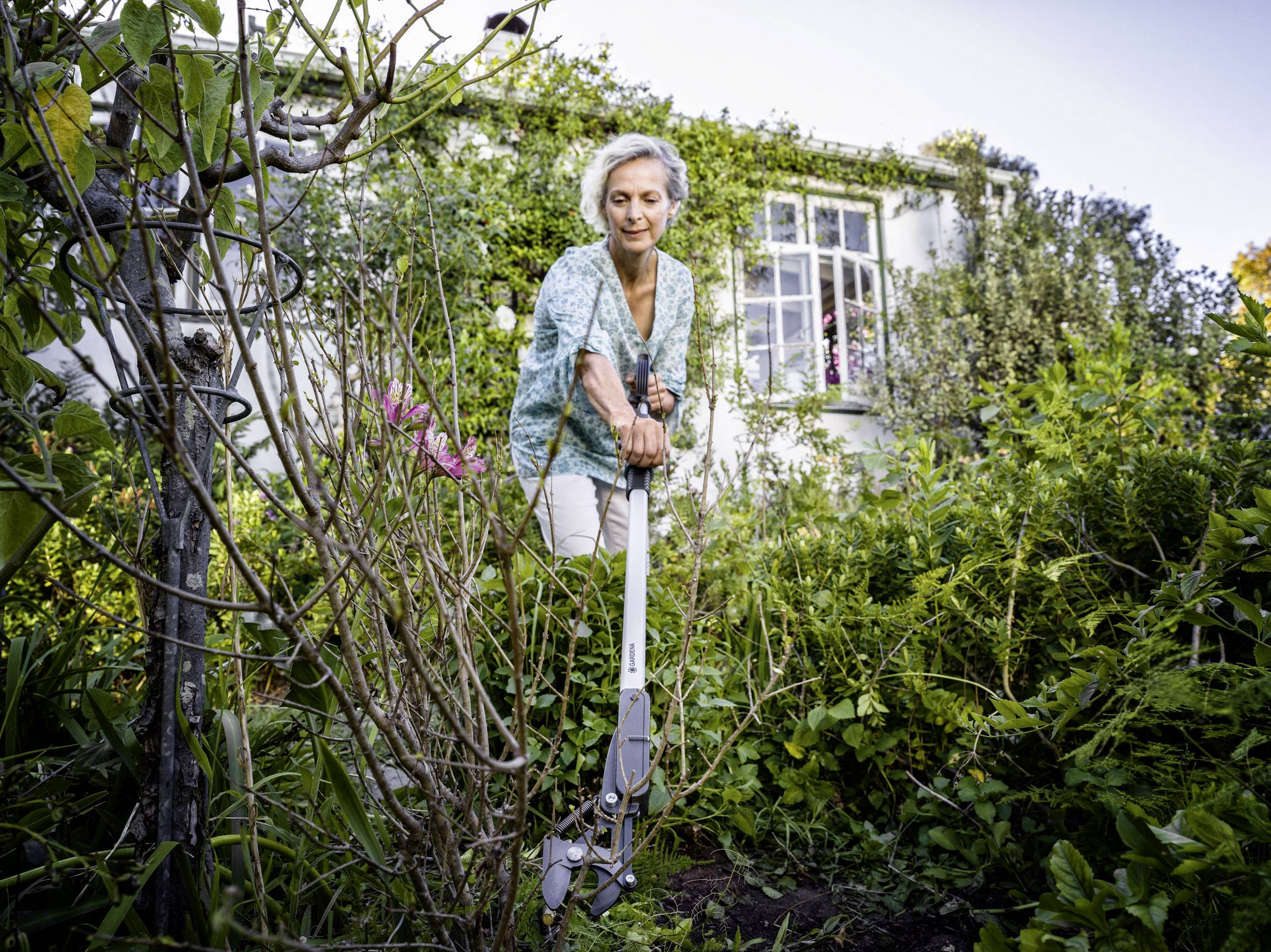 Eine Frau mittleren Alters schneidet Pflanzen in einem grünen Garten vor einem Haus. Sie trägt eine grüne Bluse und weiße Hose.