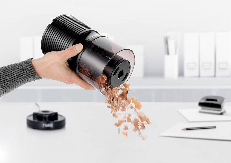 Eine Hand leert einen Staubsaugerbehälter mit Schmutz und Staub in einen Mülleimer. Bürogegenstände sind im Hintergrund sichtbar.