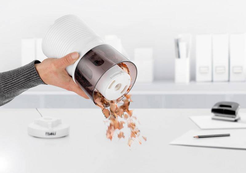 Eine Person leert einen mit Staub gefüllten Staubbehälter über einem Tisch. Im Hintergrund sind Büromaterialien zu sehen.