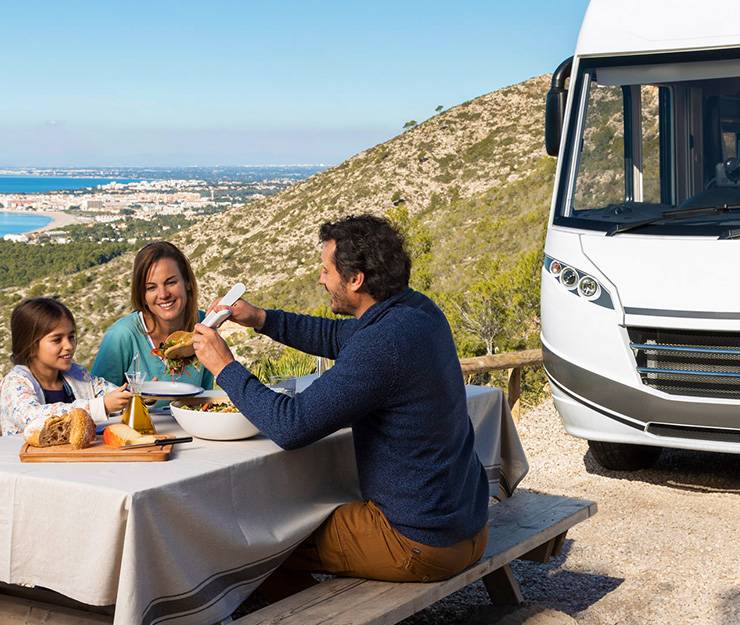 Das Bild zeigt eine Familie auf einen Rastplatz am Meer beim Mittagessen. ImHintergrund steht ein Campingmobil.