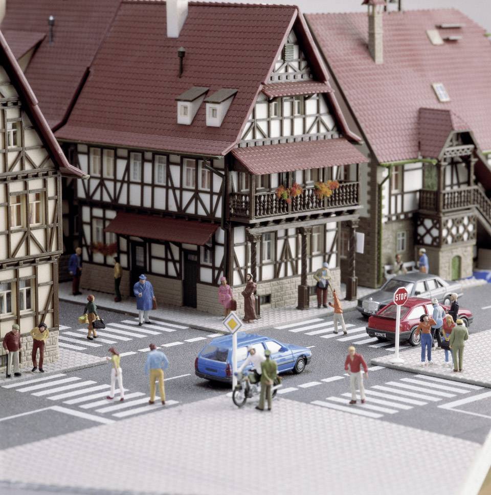 Straßenszene eines Miniaturdorfes mit Fachwerkhäusern, Autos und Figuren, die über Zebrastreifen gehen. Verkehrsschilder sind sichtbar.