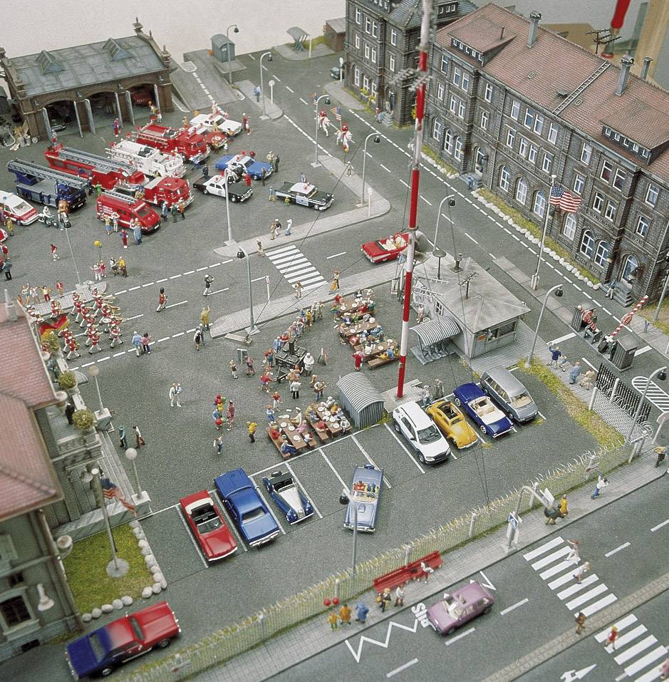 Stadtmodell mit Feuerwehrübung: Zahlreiche Miniaturfiguren, Feuerwehrautos und Zuschauer vor Stadthäusern. Kreuzung und Parkplätze sichtbar.