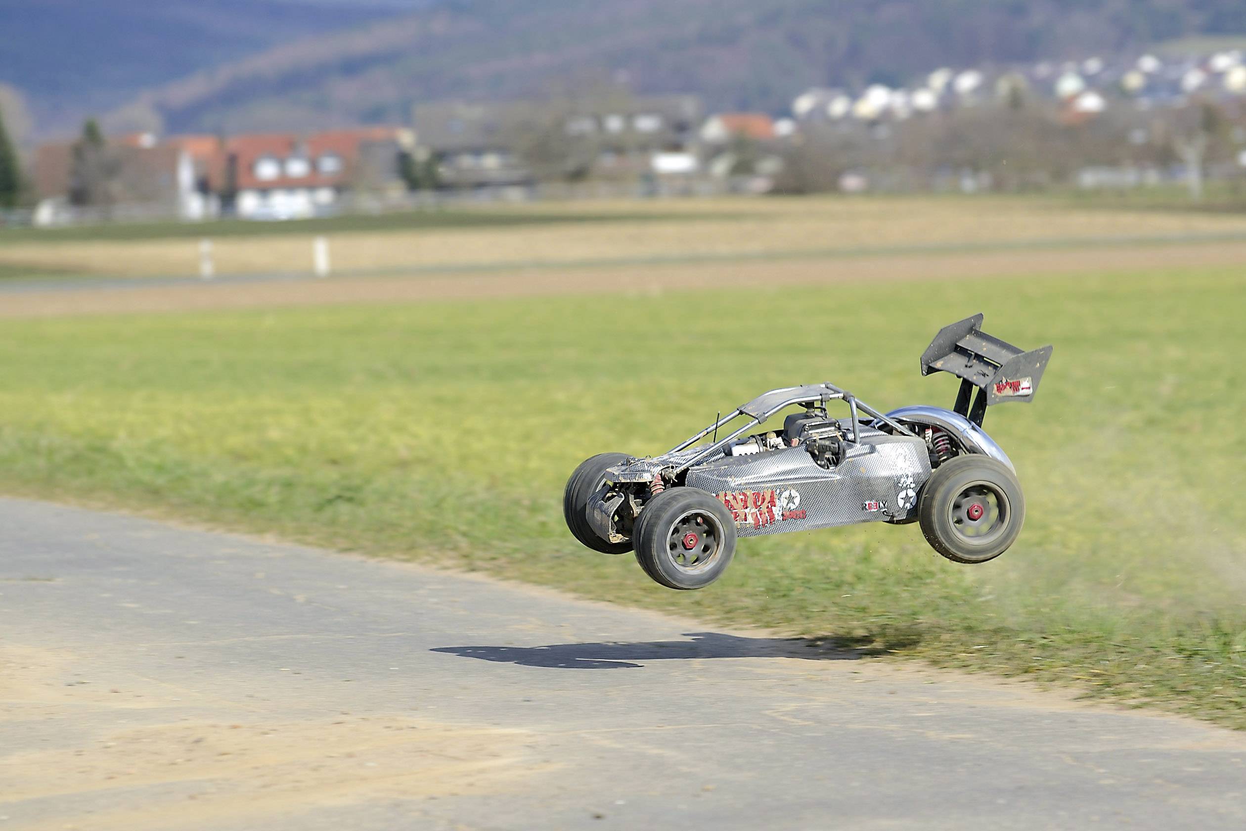 Ein ferngesteuertes Auto springt über einen gepflasterten Weg in einer ländlichen Landschaft mit Wiesen und Hügeln im Hintergrund.