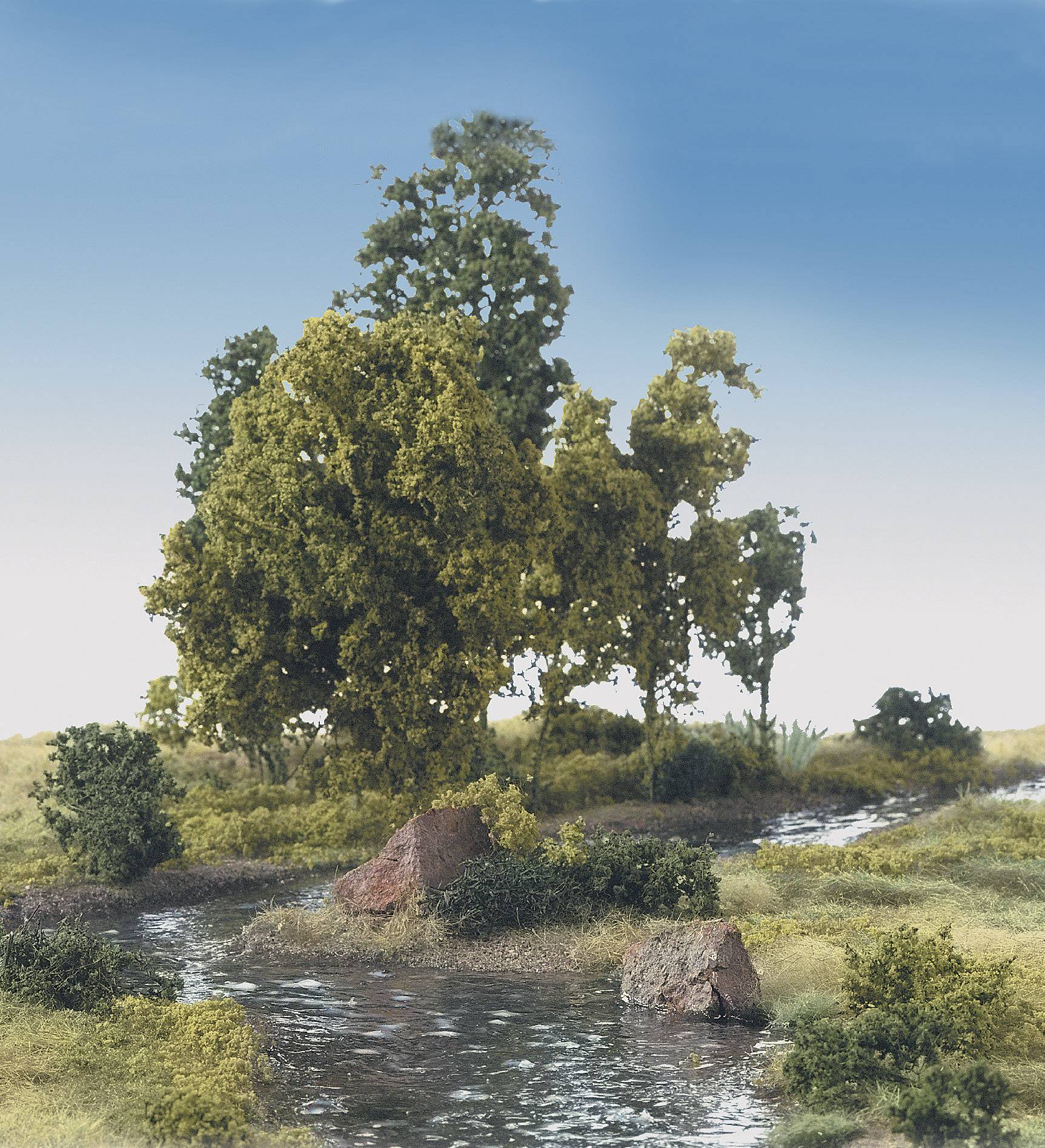 Eine kleine Landschaft mit Bach, umgeben von Bäumen und Sträuchern unter blauem Himmel.