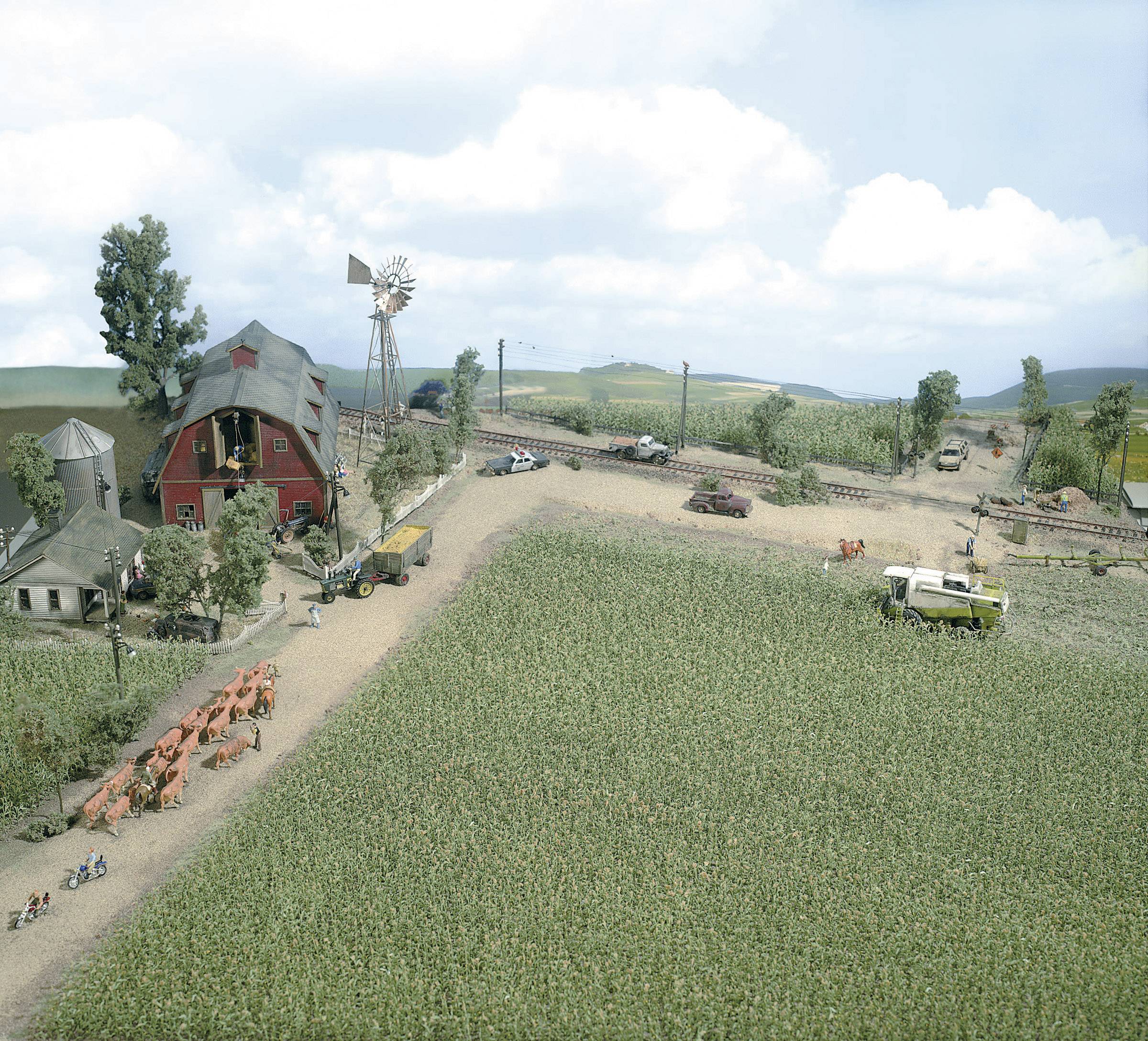 Luftbild eines Bauernhofs mit einem roten Scheunenhaus, Windmühle und Feldern. Kühe grasen, und landwirtschaftliche Maschinen sind sichtbar.