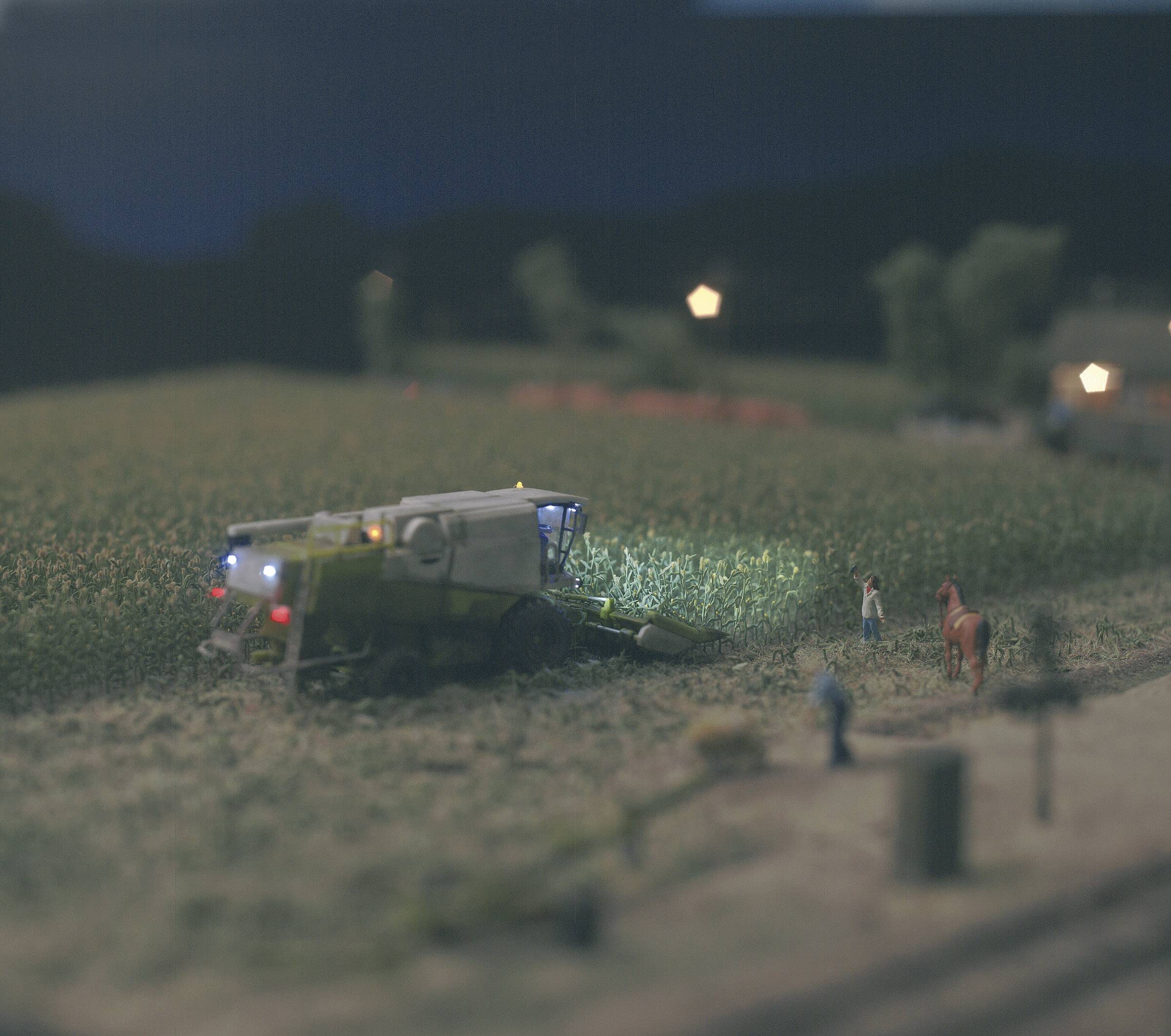Ein Mähdrescher arbeitet nachts auf einem Feld. Zwei Personen stehen am Rand, neben ihnen ein Pferd. Im Hintergrund sind Lichter zu sehen.