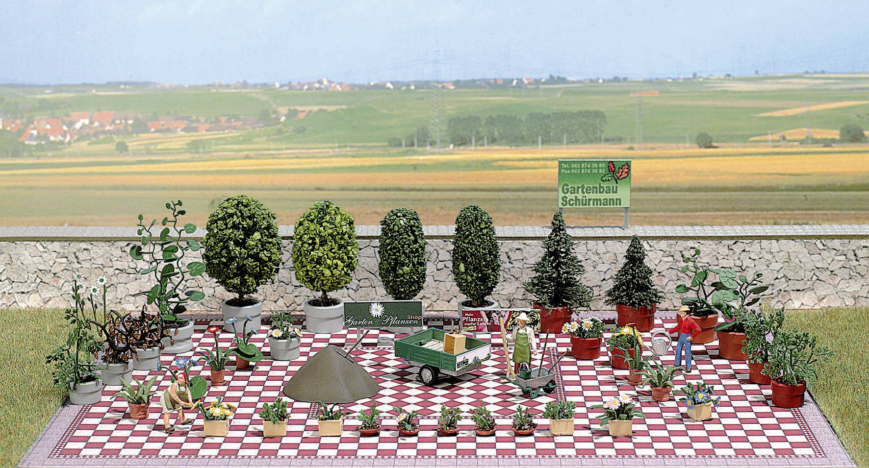 Modell-Landschaft mit Miniatur-Bäumen und Pflanzen auf einem Schachbrettmusterboden. Im Hintergrund weite Felder und ein Schild.