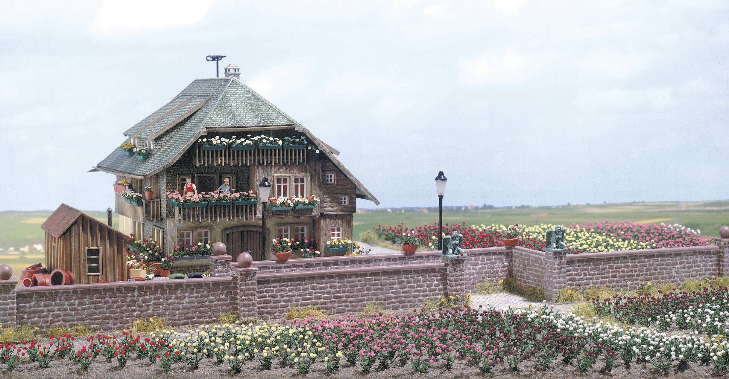 Ein ländliches Haus mit Holzbalkon und blühenden Blumenbeeten im Vordergrund, umgeben von einer Backsteinmauer, vor einem weiten Feld.