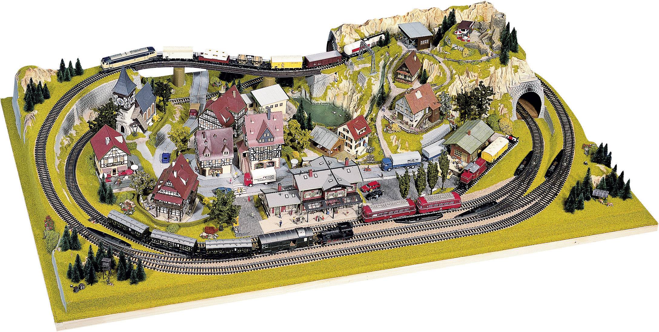 Modelleisenbahnlandschaft mit mehreren Zügen, die auf Gleisen durch ein Dorf mit Gebäuden und Bäumen fahren, umgeben von Landschaft.