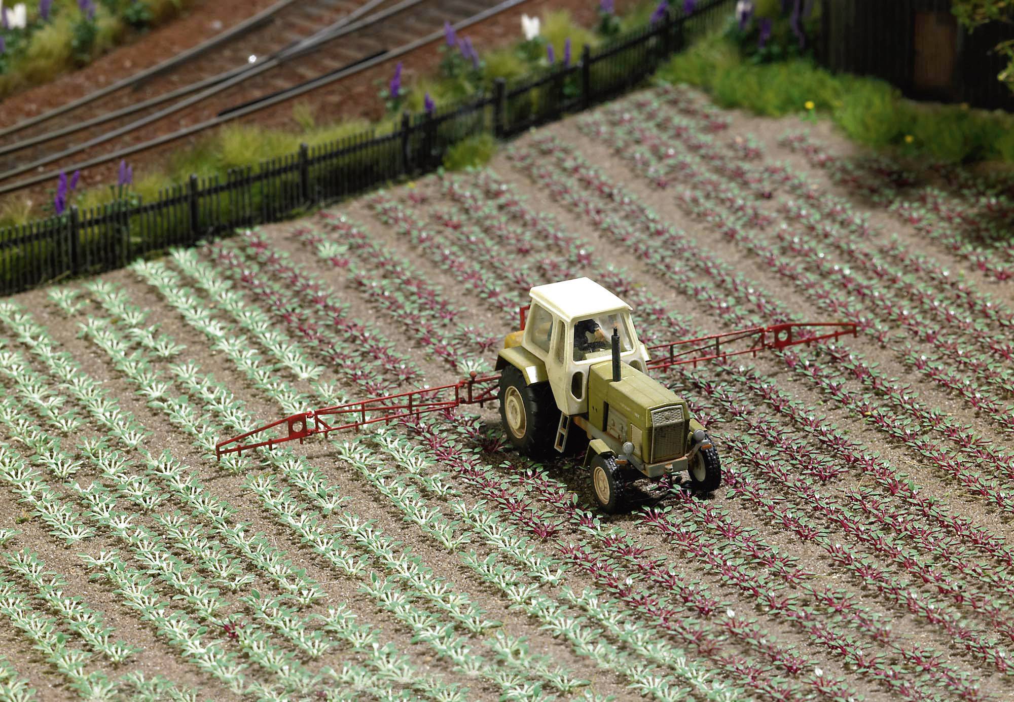 Ein kleiner Traktor fährt über ein Miniaturfeld und sprüht Pestizide auf Pflanzen. Ein Bahngleis verläuft im Hintergrund.