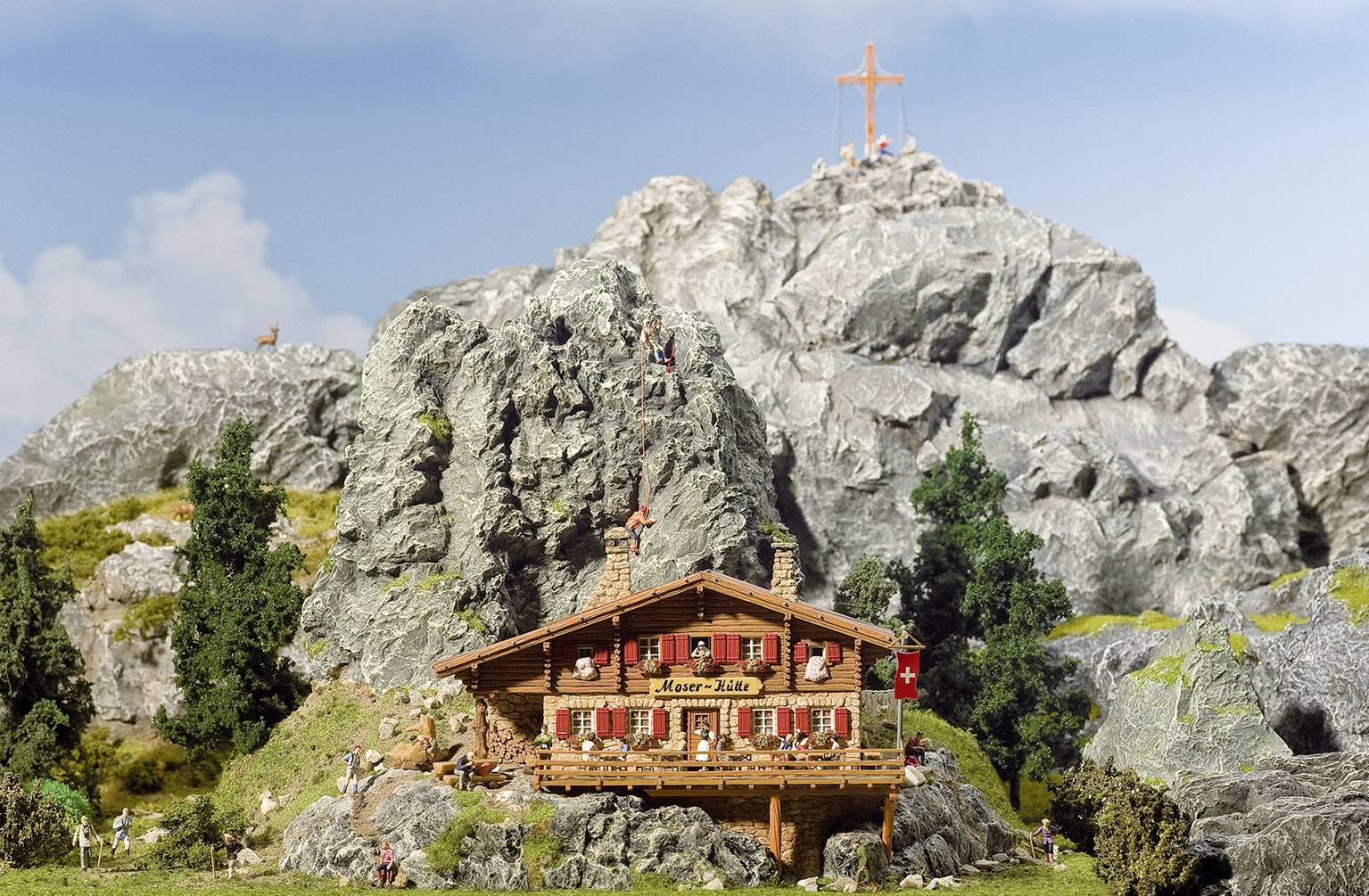 Ein Holzhaus in den Bergen, umgeben von Felsen und Bäumen, mit einem Kreuz auf dem Berggipfel. Menschen wandern und klettern in der Umgebung.