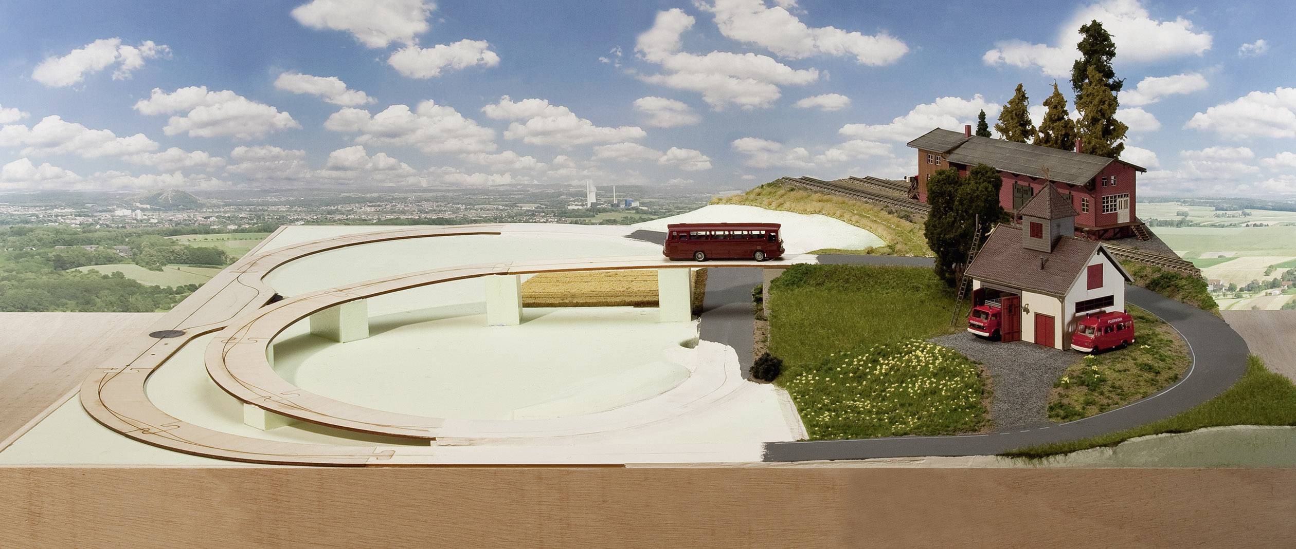 Miniaturmodell eines Landschaftsdioramas mit Straße, Bus und zwei Häusern. Im Hintergrund eine weite Landschaft unter einem blauen Himmel.