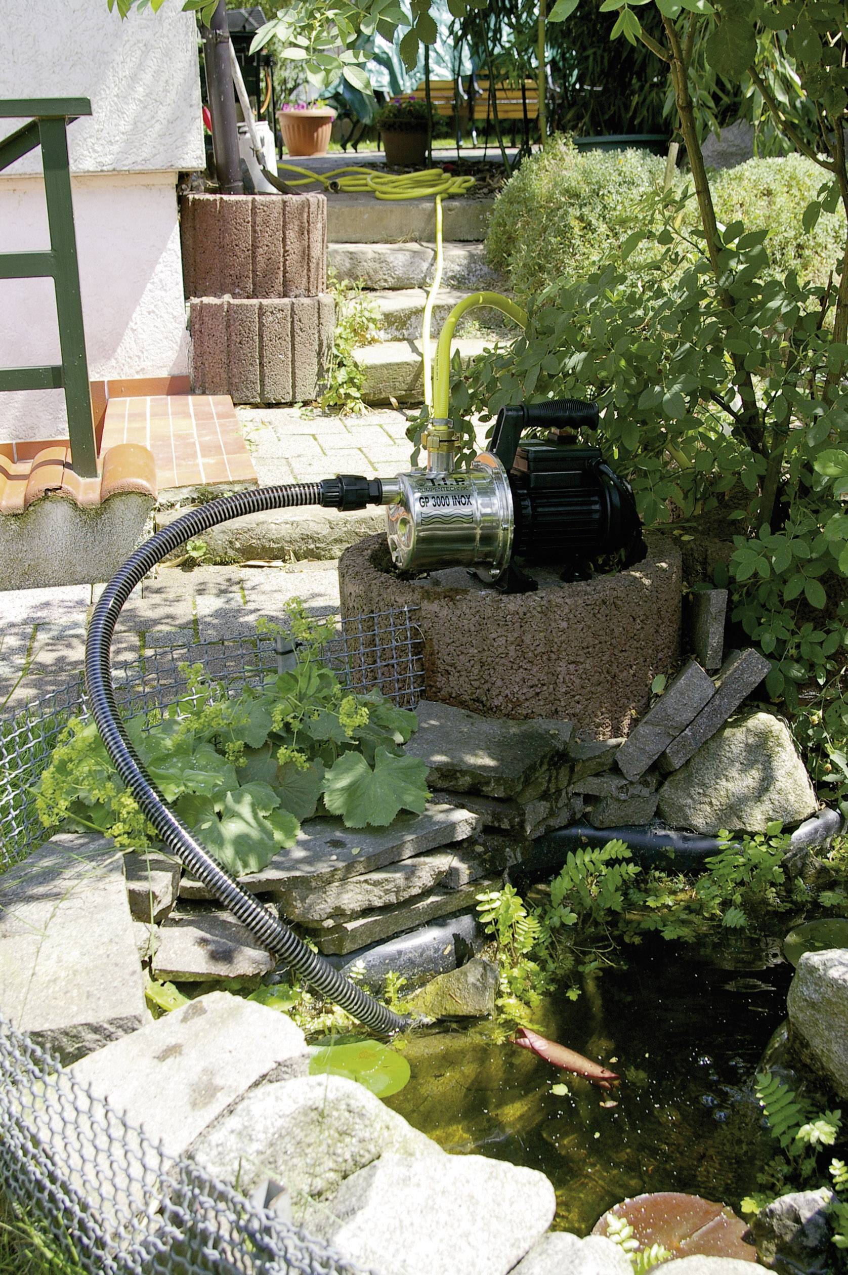 Gartenpumpe am Teich platziert, umgeben von Pflanzen und Deko-Steinen. Rohr verbindet Pumpe mit Wasser. Im Wasser schwimmt ein Fisch.