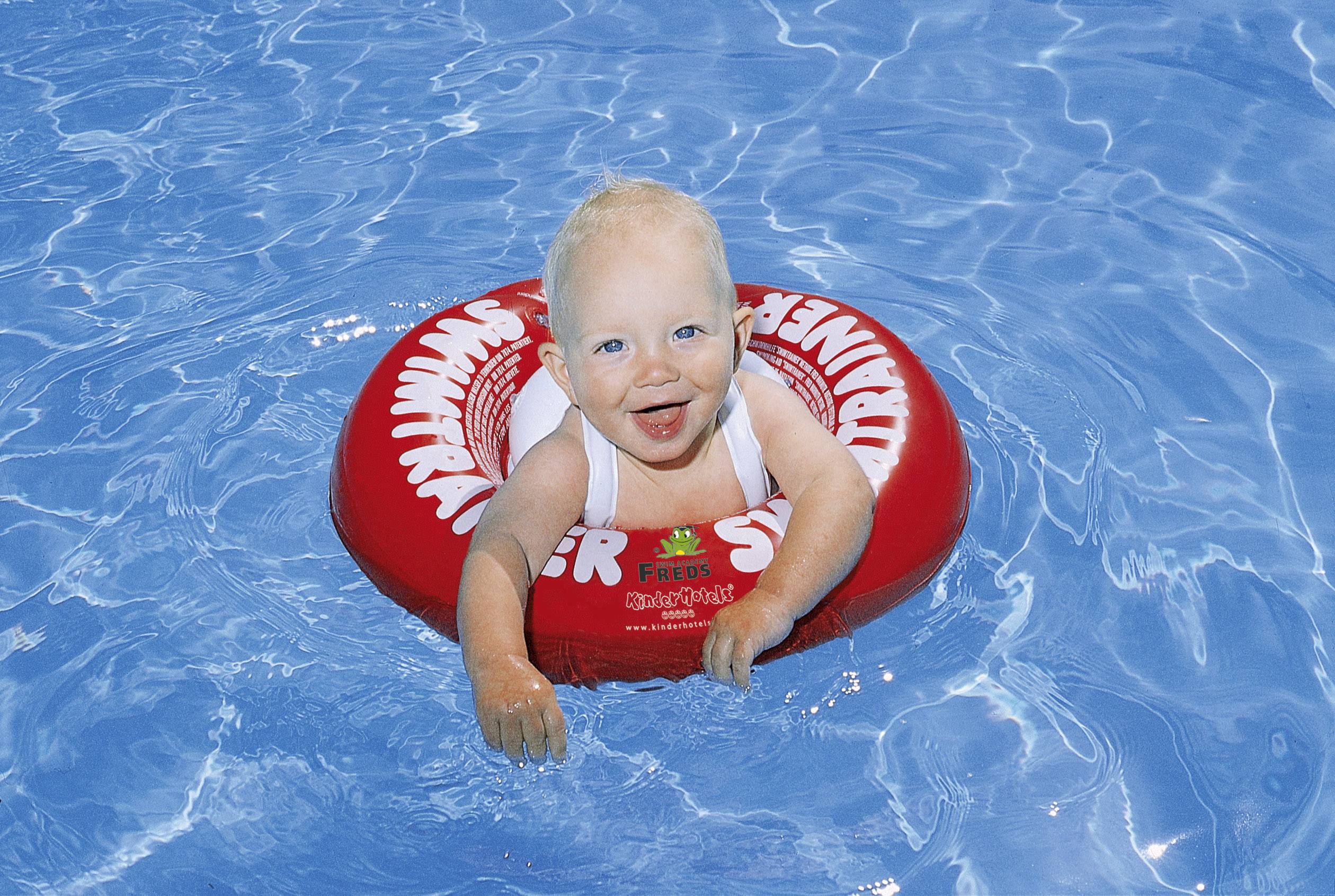 Kleinkind lächelnd in einem roten Schwimmreifen mit Aufschrift im blauen Poolwasser.