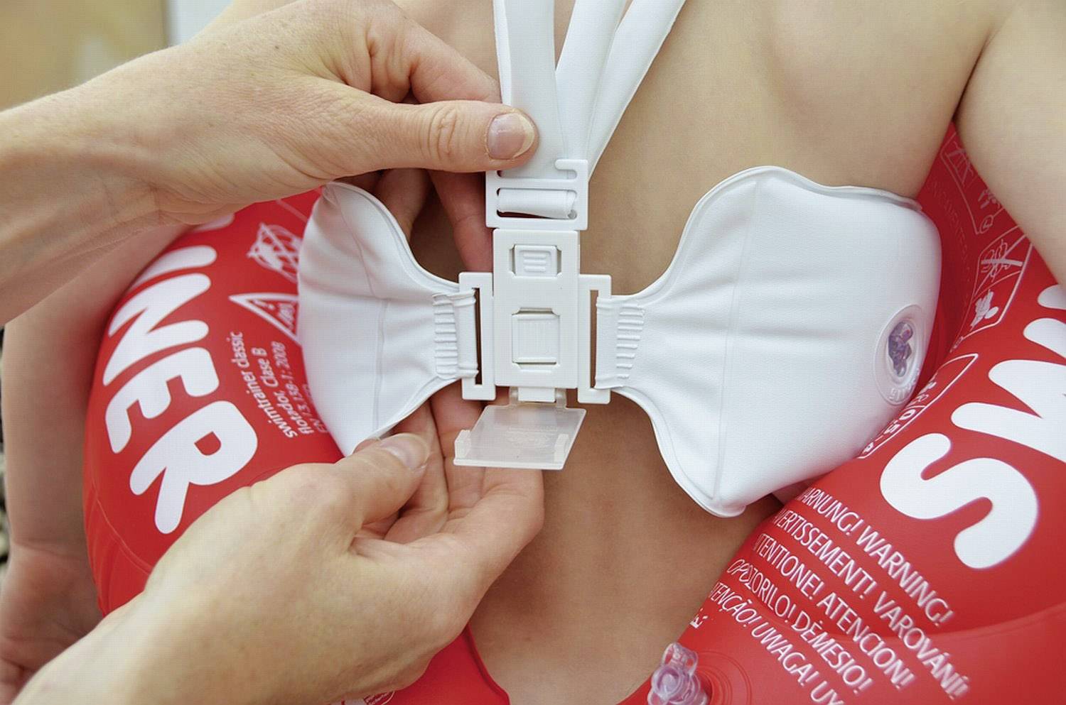 Eine Person befestigt einen weißen Sicherheitsverschluss an der Schwimmweste eines Kindes. Der Hintergrund zeigt Teile der roten Weste mit Aufschrift.