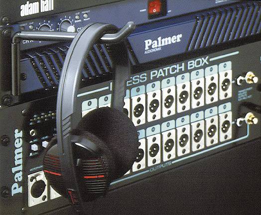 Ein Kopfhörer hängt an einer Audiogeräte-Rack mit mehreren Ein- und Ausgängen. Auf dem Rack sind die Marken 'Palmer' und 'adam hall' sichtbar.