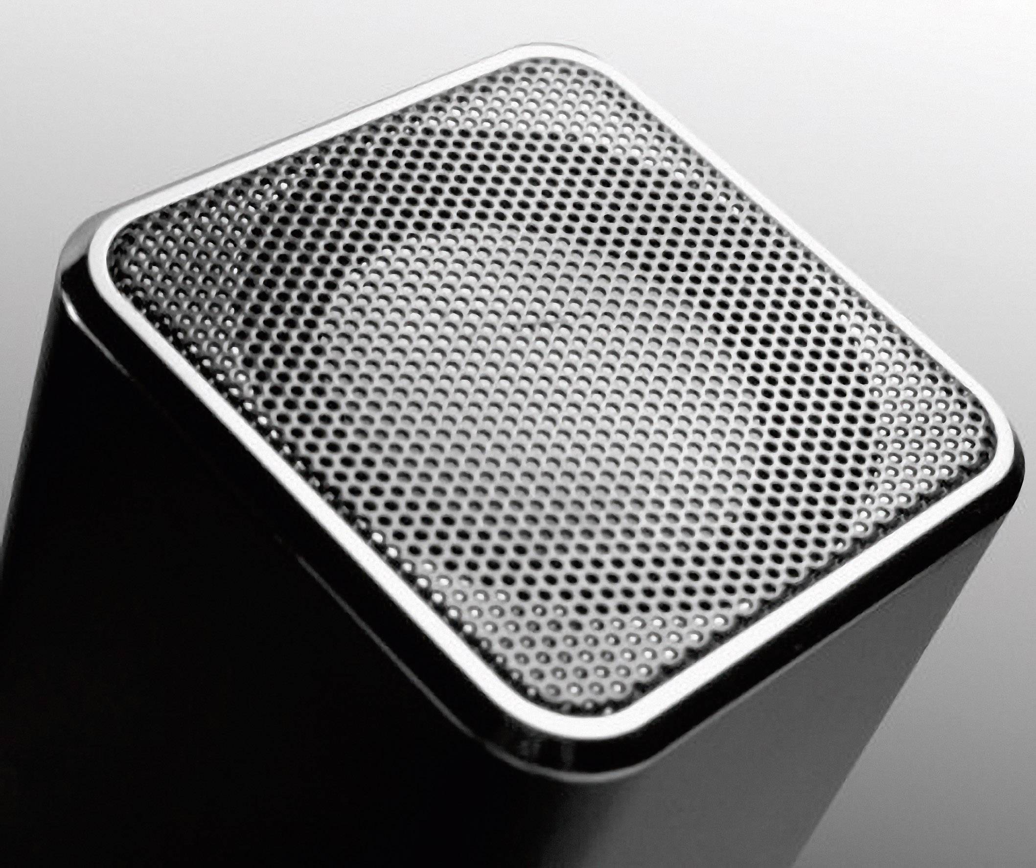 Ein quadratischer Lautsprecher mit metallischem Gitter vor grauem Hintergrund.