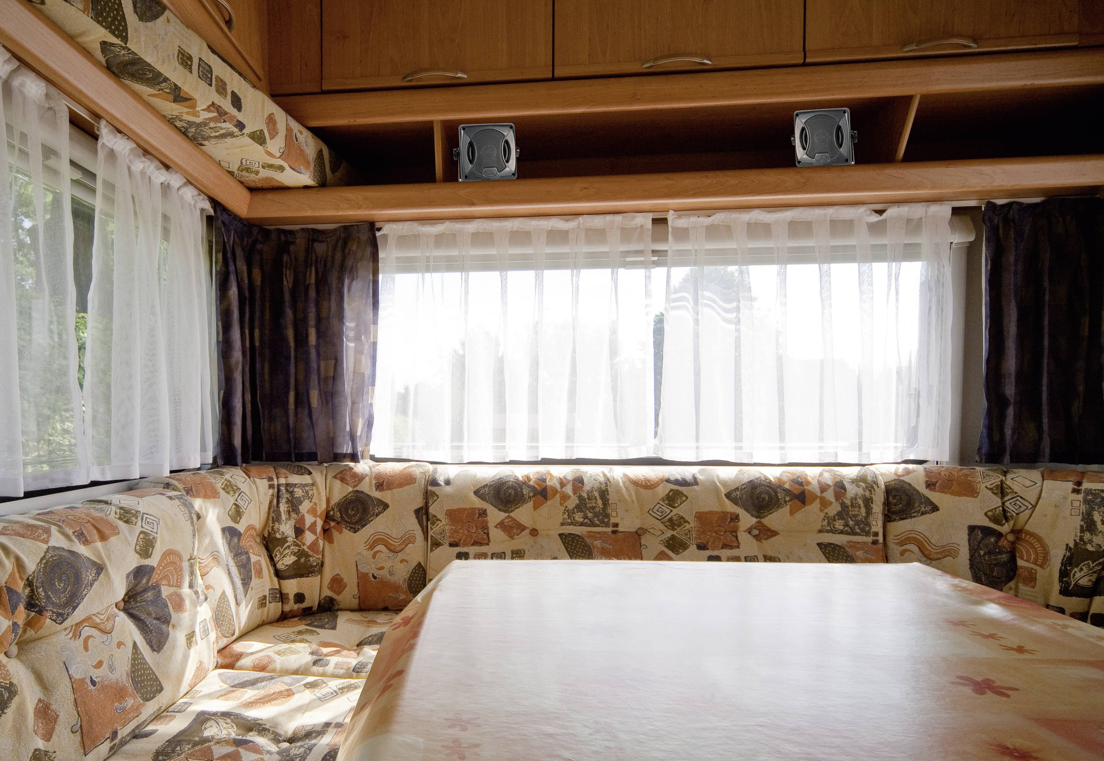 Wohnmobil-Innenraum mit gepolsterter Eckbank und Esstisch vor einem großen Fenster mit Vorhängen. Hölzerne Schränke sind oben zu sehen.