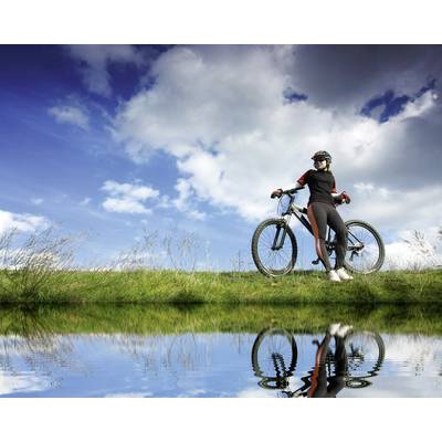 Eine Person steht mit Fahrrad am Ufer eines Gewässers unter blauem Himmel mit Wolken. Das Bild spiegelt sich im Wasser.