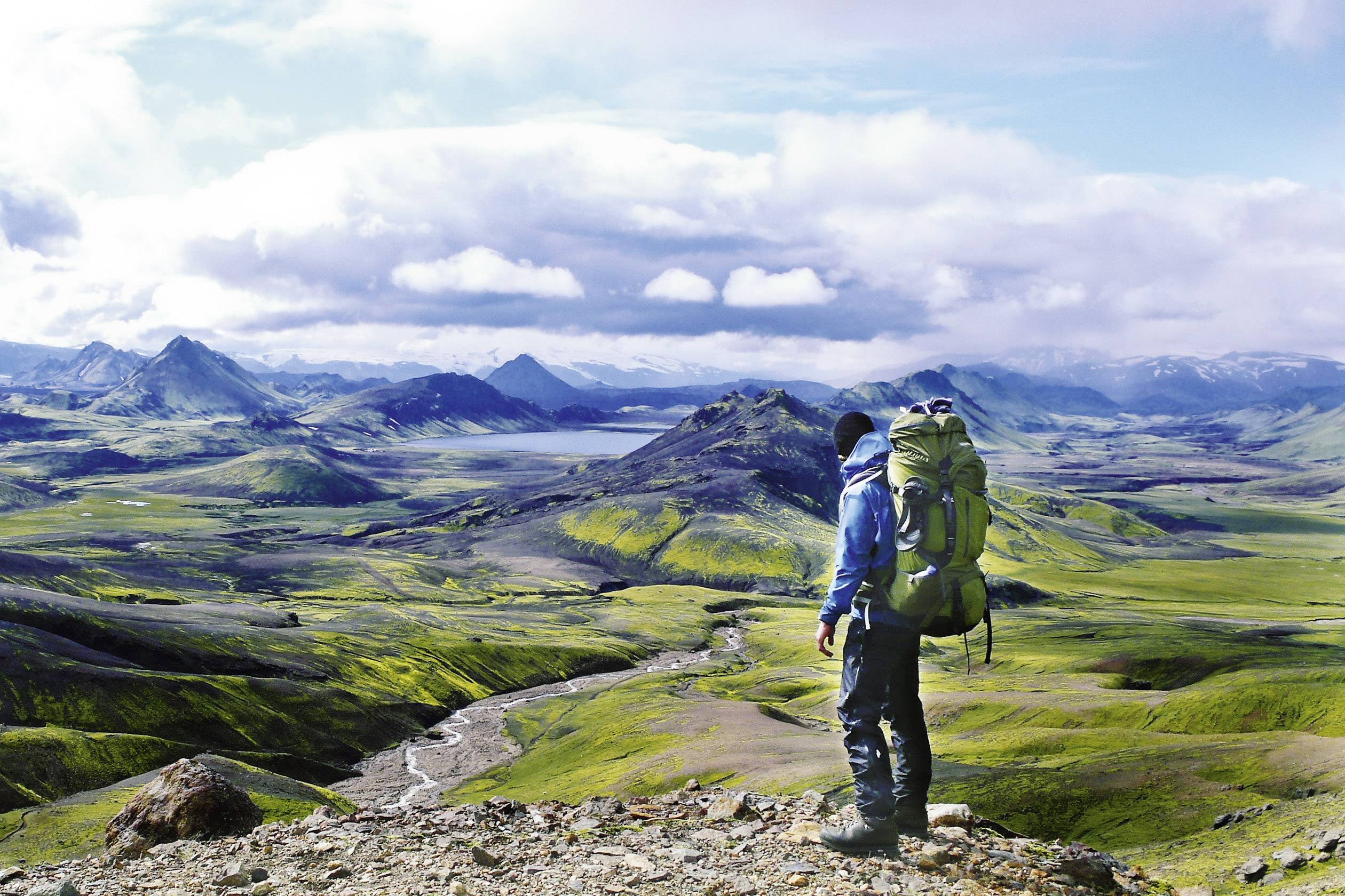 Eine Person steht mit einem großen Rucksack auf einem Hügel und blickt auf eine weite, grüne Landschaft mit Bergen und Wolken am Horizont.