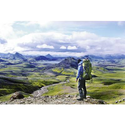 Eine Person steht mit einem großen Rucksack auf einem Hügel und blickt auf eine weite, grüne Landschaft mit Bergen und Wolken am Horizont.