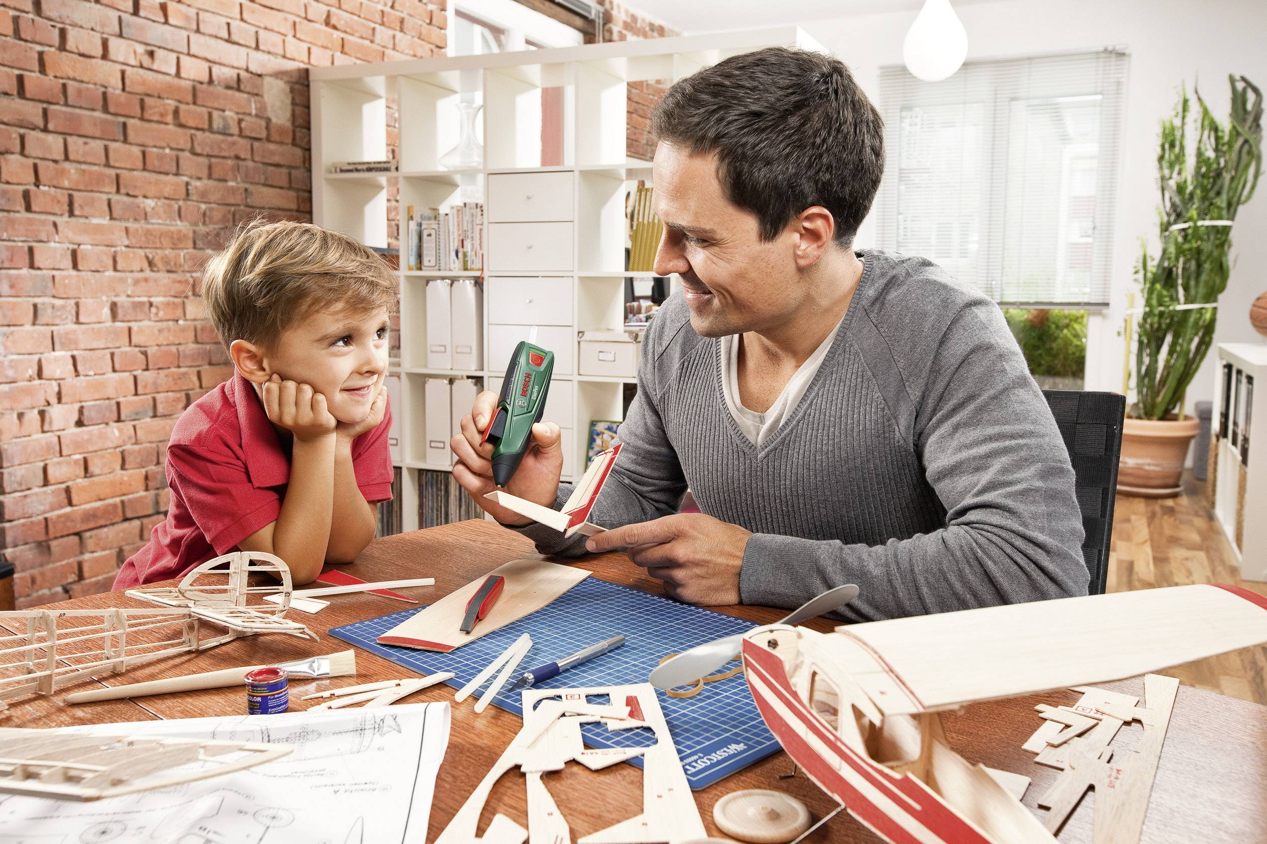 Ein Mann und ein Junge bauen Modellflugzeuge aus Holz am Tisch. Der Junge schaut interessiert zu, während der Mann ein Stück zeigt.