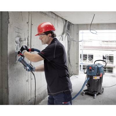 Ein Bauarbeiter trägt einen roten Schutzhelm und benutzt eine Bohrmaschine an einer Betonwand. Ein Industriesauger steht daneben.