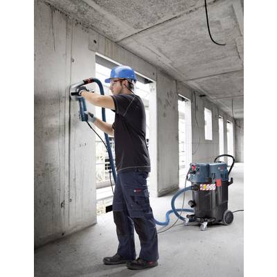 Eine Person mit Schutzhelm arbeitet mit einem Bohrhammer an einer Betonwand in einem halbfertigen Gebäude. Ein Industriesauger steht daneben.