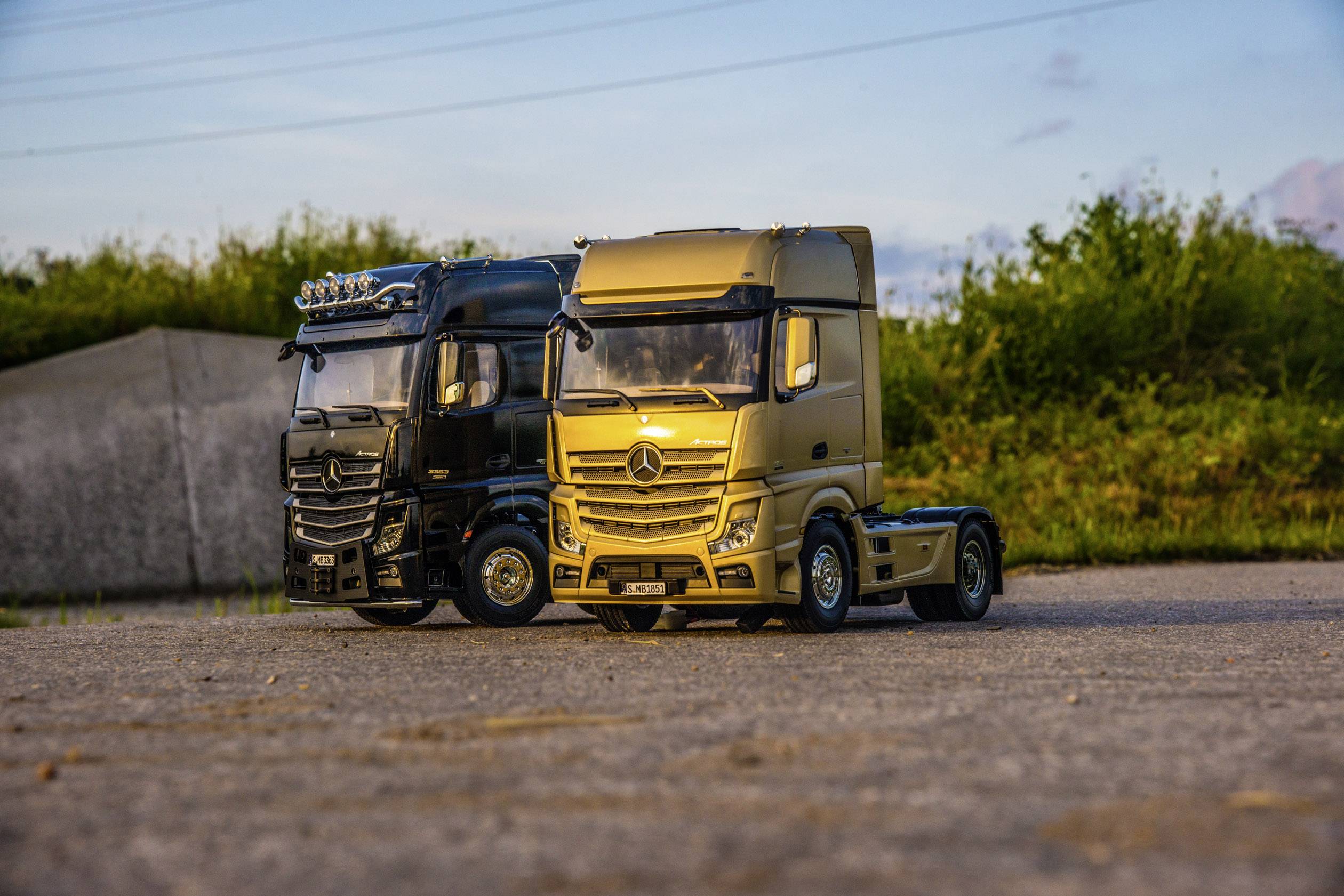 Zwei Modell-LKWs auf einer Straße, einer in Schwarz und einer in Gold. Im Hintergrund sind grüne Büsche und der Himmel sichtbar.