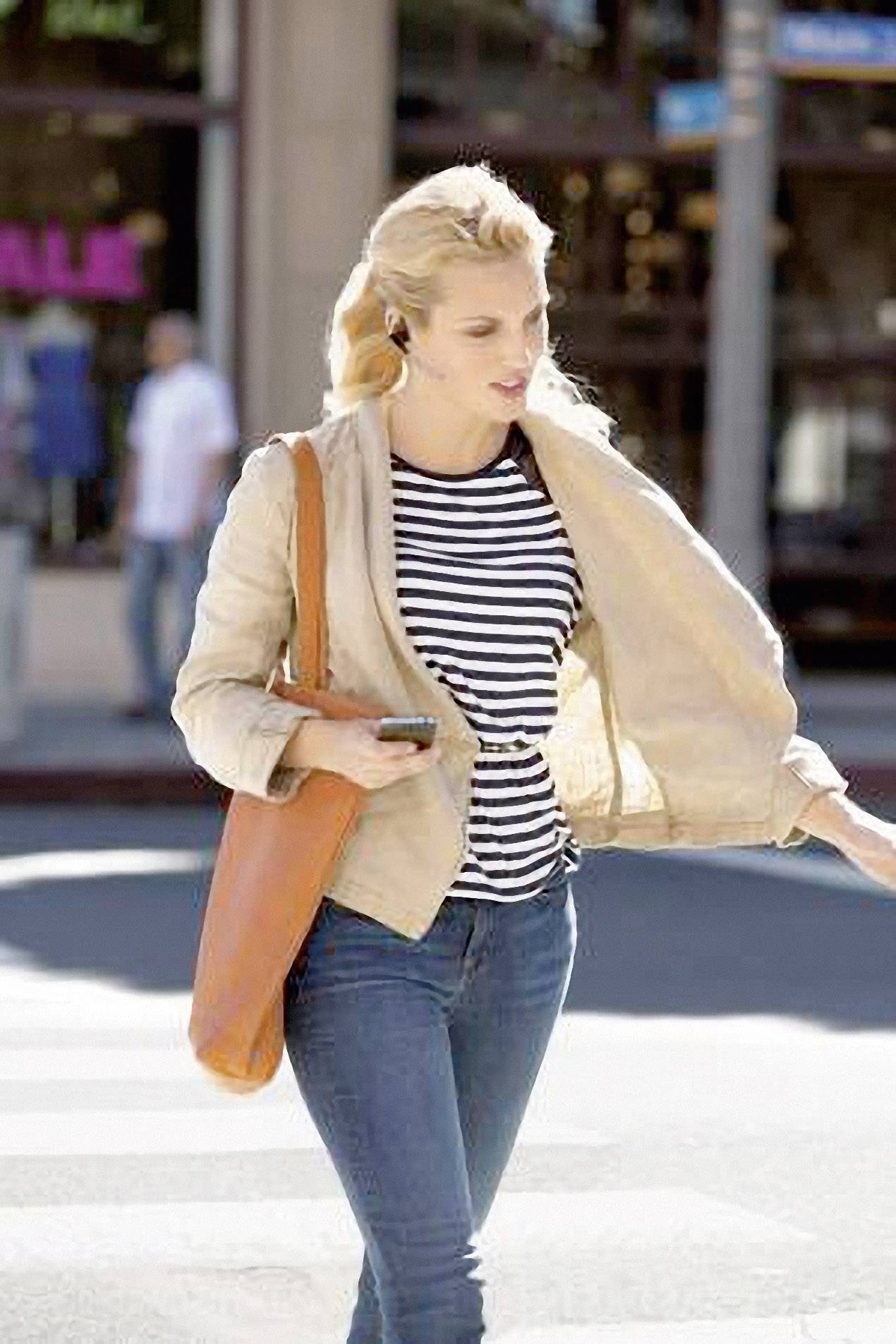 Eine Frau überquert die Straße bei Tageslicht. Sie trägt eine gestreifte Bluse, eine beige Jacke, blaue Jeans und hat eine orangene Tasche.