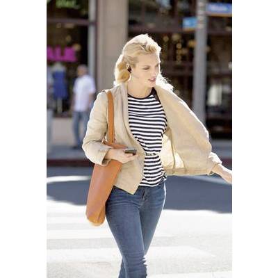 Eine Frau überquert die Straße bei Tageslicht. Sie trägt eine gestreifte Bluse, eine beige Jacke, blaue Jeans und hat eine orangene Tasche.
