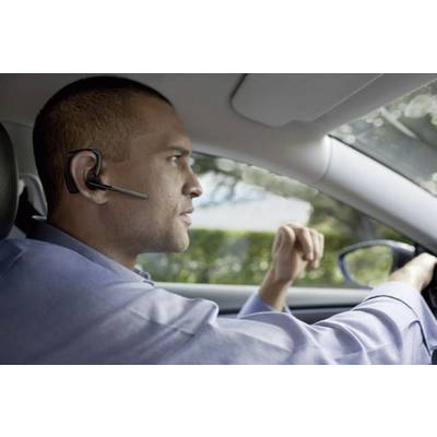 Ein Mann fährt ein Auto und trägt ein Headset, während er konzentriert blickt. Die Fahrt scheint bei Tageslicht zu erfolgen.