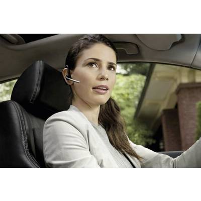 Eine Frau sitzt in einem Auto und spricht in ein Headset. Sie trägt einen hellen Blazer. Im Hintergrund ist ein Haus zu sehen.