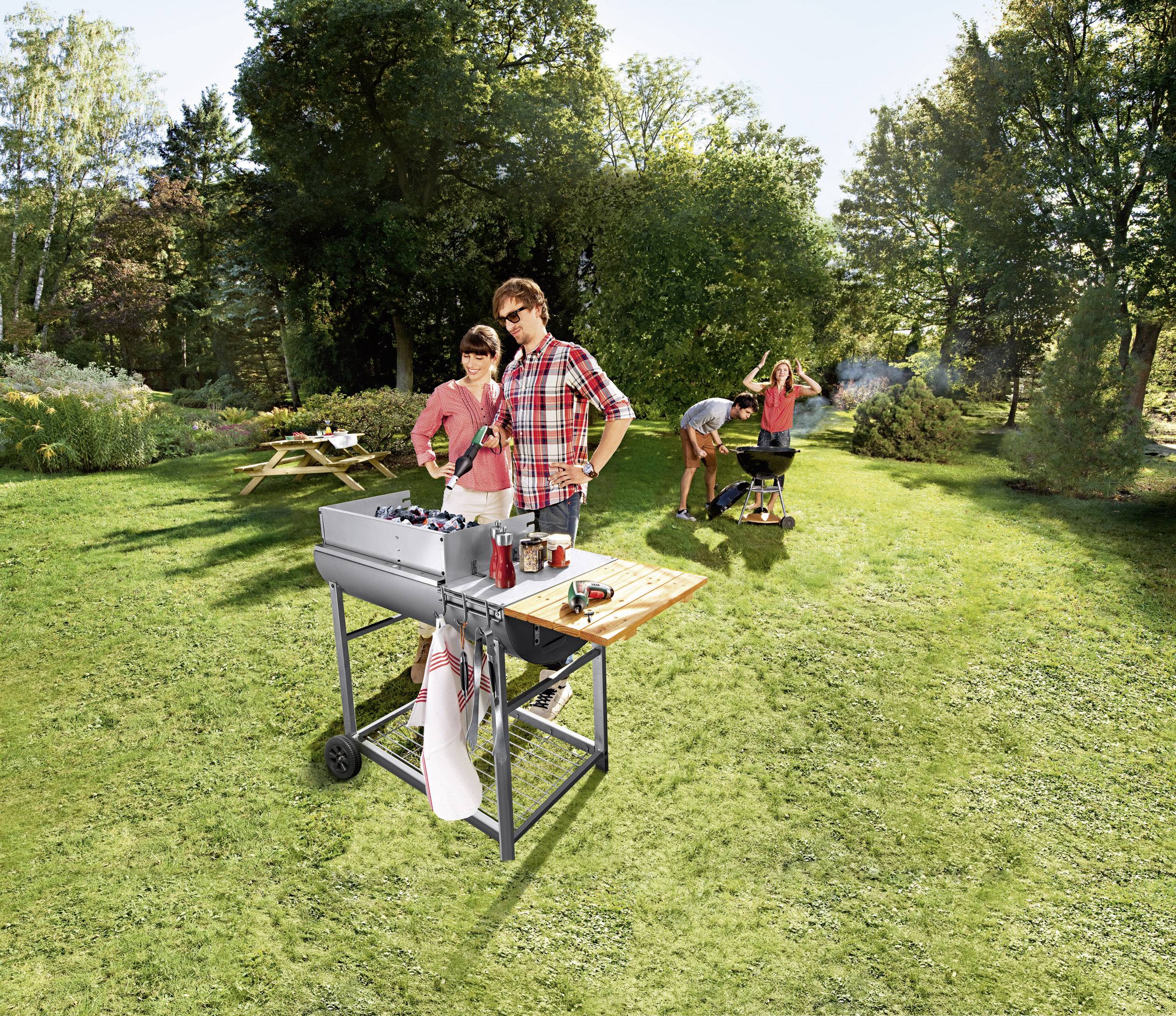 Eine Gruppe von vier Personen grillt im Freien auf einer grünen Wiese. Zwei Personen stehen am Grill und scheinen zufrieden, während eine andere Person den Grill mit Kohle anheizt.