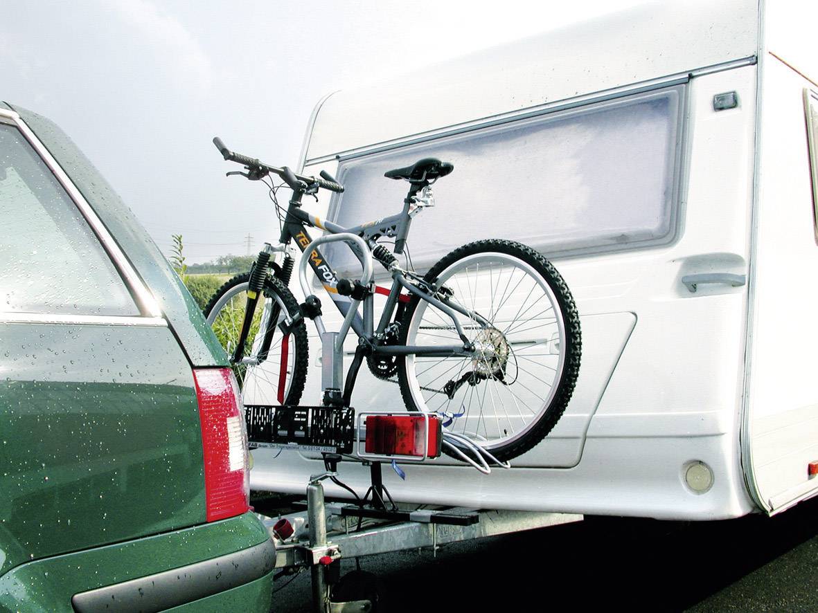 Ein grünes Auto zieht einen Wohnwagen. Ein Fahrrad ist am Heck des Wohnwagens befestigt. Es regnet leicht.