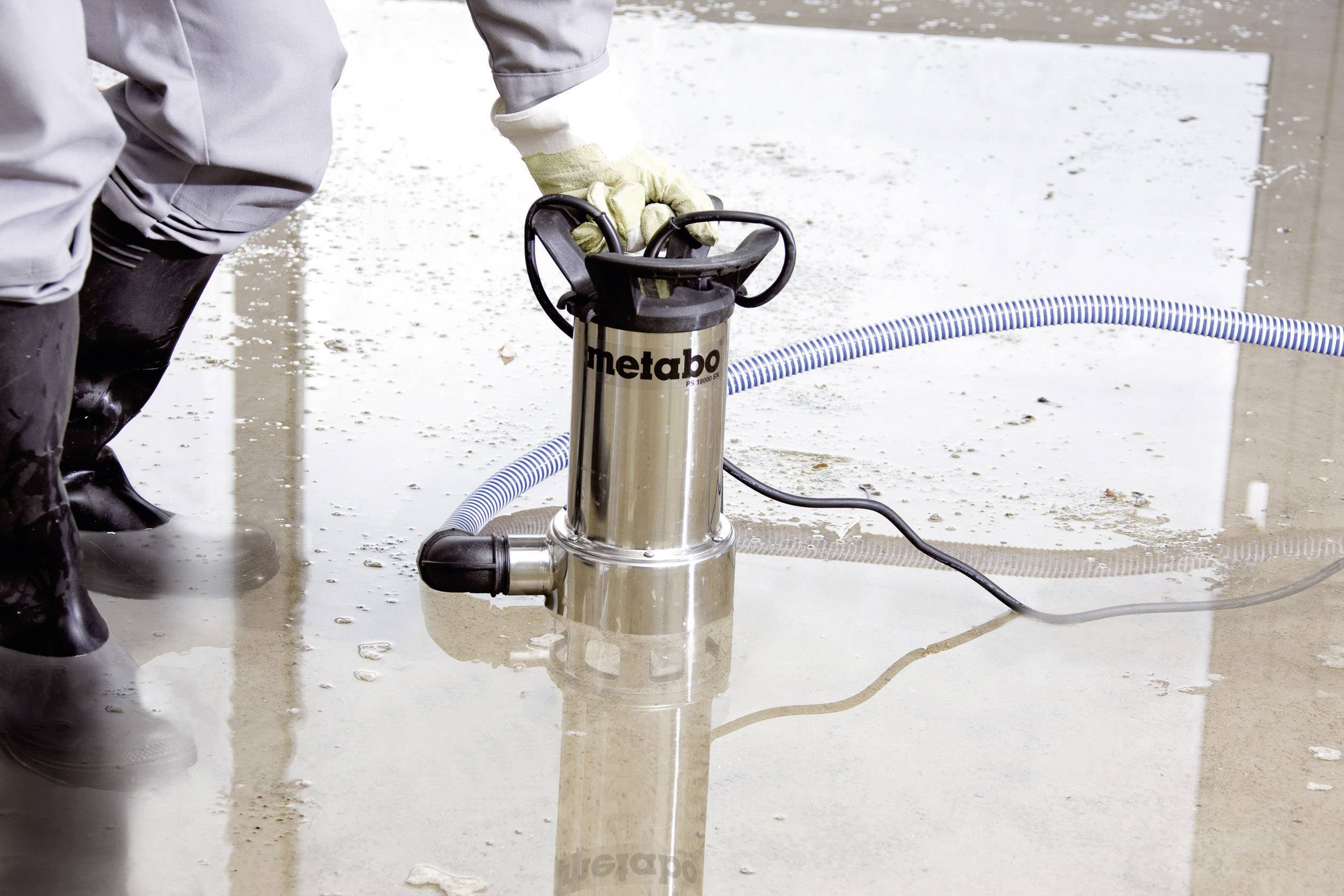 Eine Person bedient eine Tauchpumpe zum Abpumpen von Wasser auf einer überfluteten Betonfläche. Die Pumpe ist mit einem blauen Schlauch verbunden.