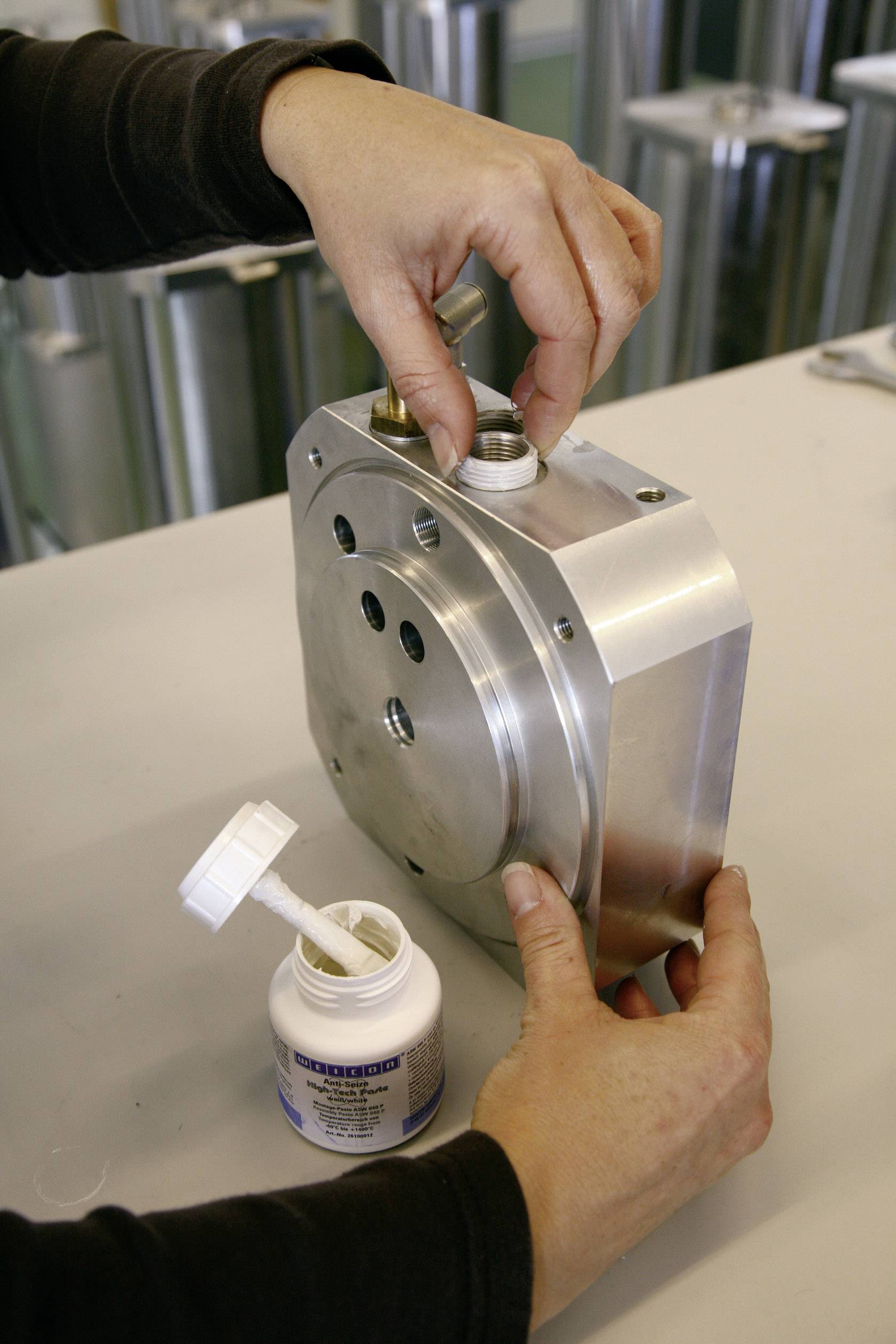 Person verbindet ein Metallteil mit Schrauben. Eine offene Flasche mit Dichtstoff steht daneben auf der Werkbank.