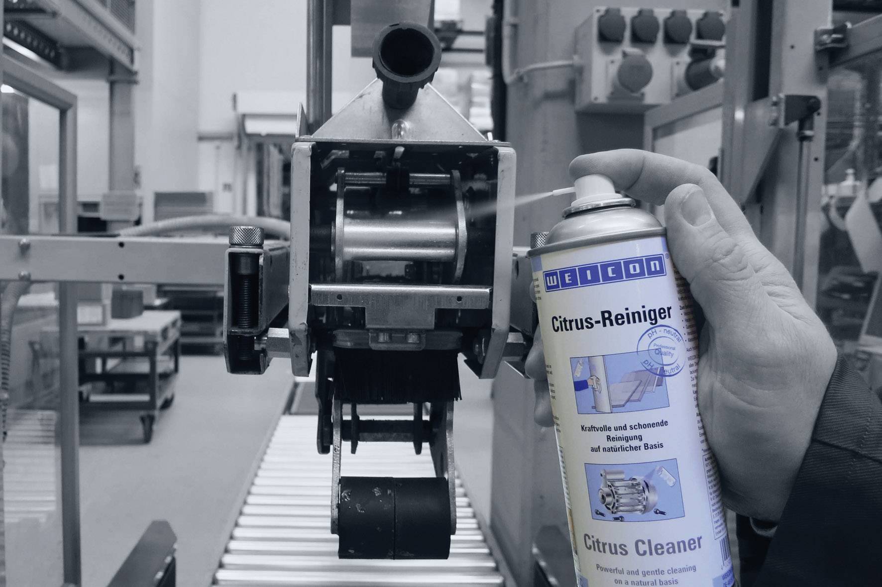 Eine Hand sprüht eine Dose 'Citrus-Reiniger' auf einen Metallteil in einer Fabrikumgebung. Reinigungsvorgang in industrieller Halle.