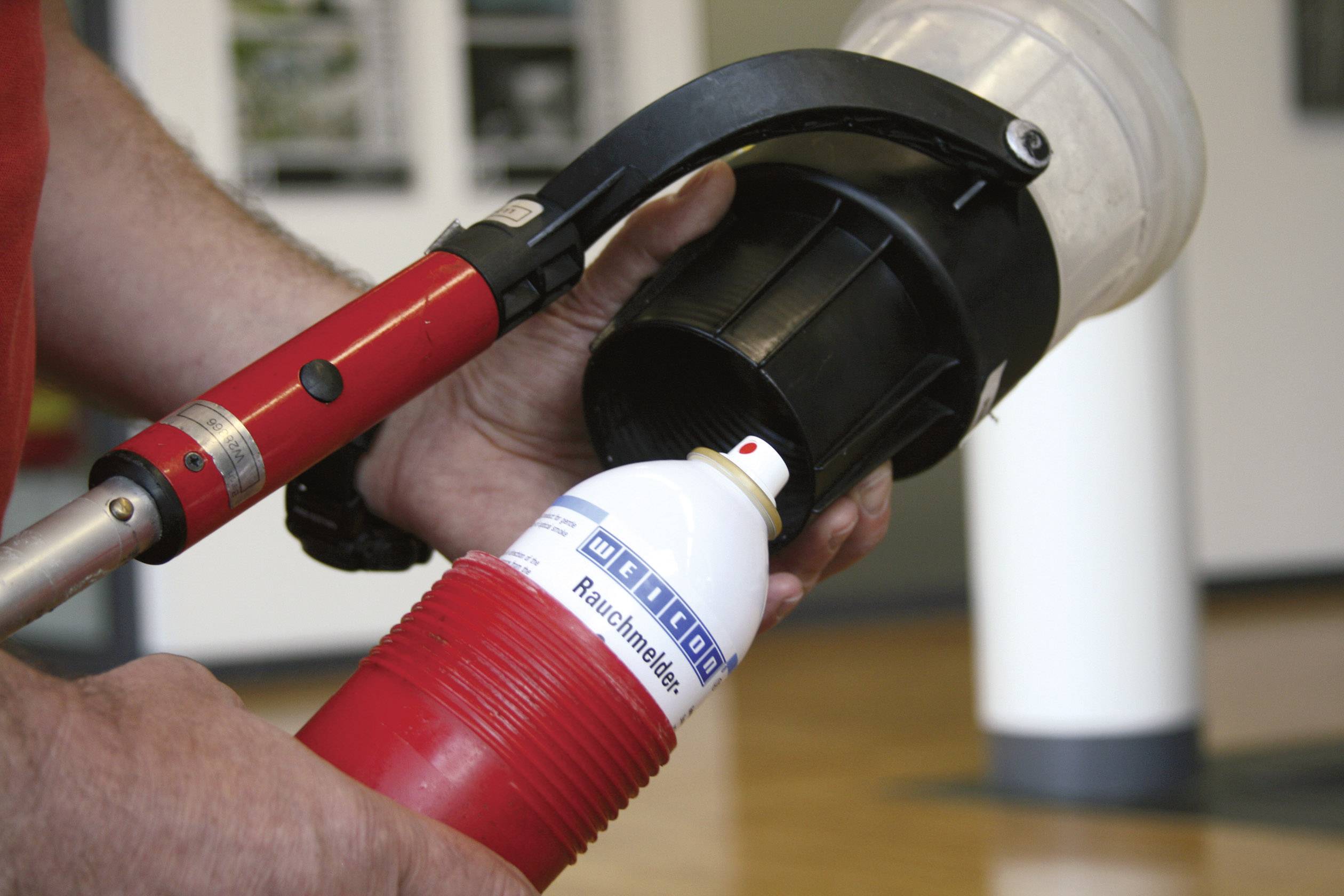 Eine Person testet einen Rauchmelder mit einer Sprühdose. Die Dose ist rot-weiß und wird gegen den Melder gedrückt.