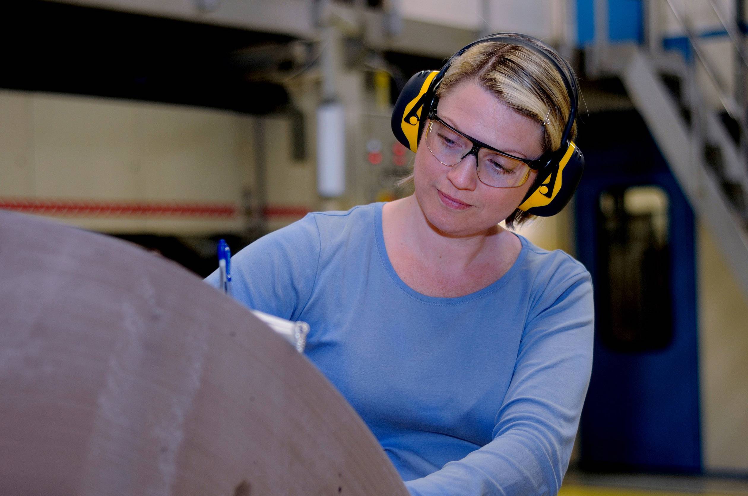 Eine Frau mit Gehörschutz und Schutzbrille schreibt Notizen in einer Industriewerkstatt. Sie trägt ein blaues Shirt.