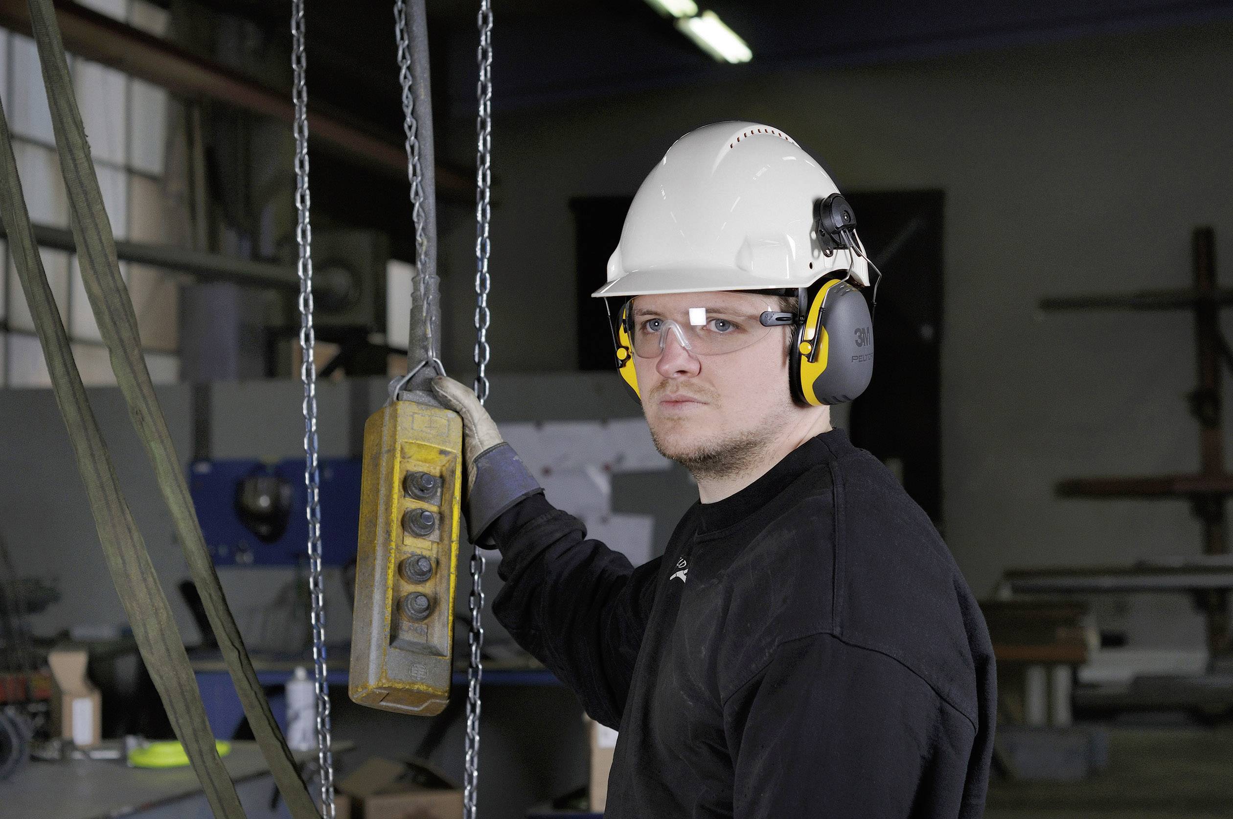 Ein Arbeiter trägt Schutzhelm und Gehörschutz, hält eine Kettensteuerung in der Hand in einer industriellen Umgebung.