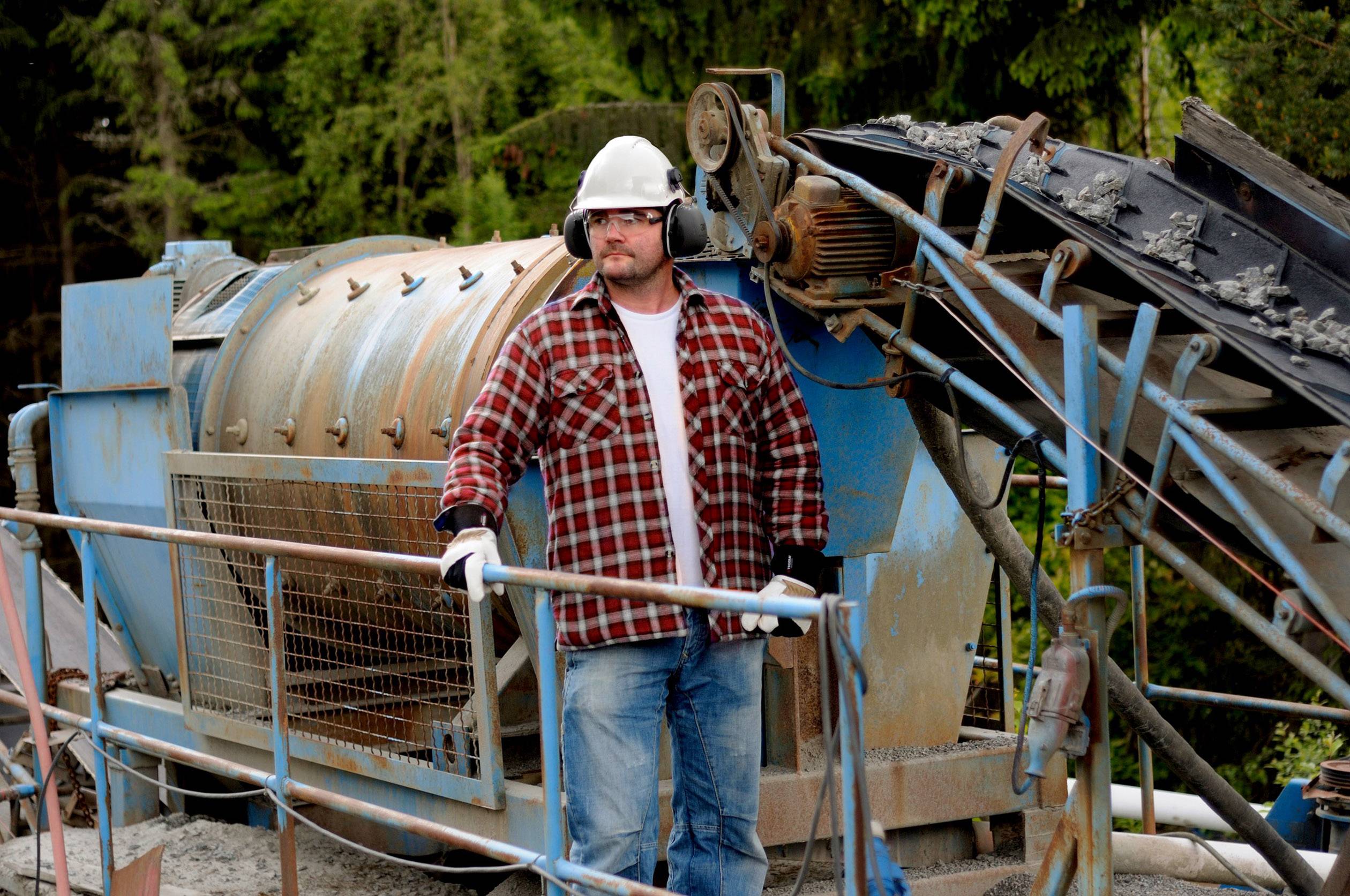 Ein Arbeiter in Schutzkleidung steht vor einer industriellen Maschine in einem Außenbereich. Die Umgebung ist von Bäumen umgeben.