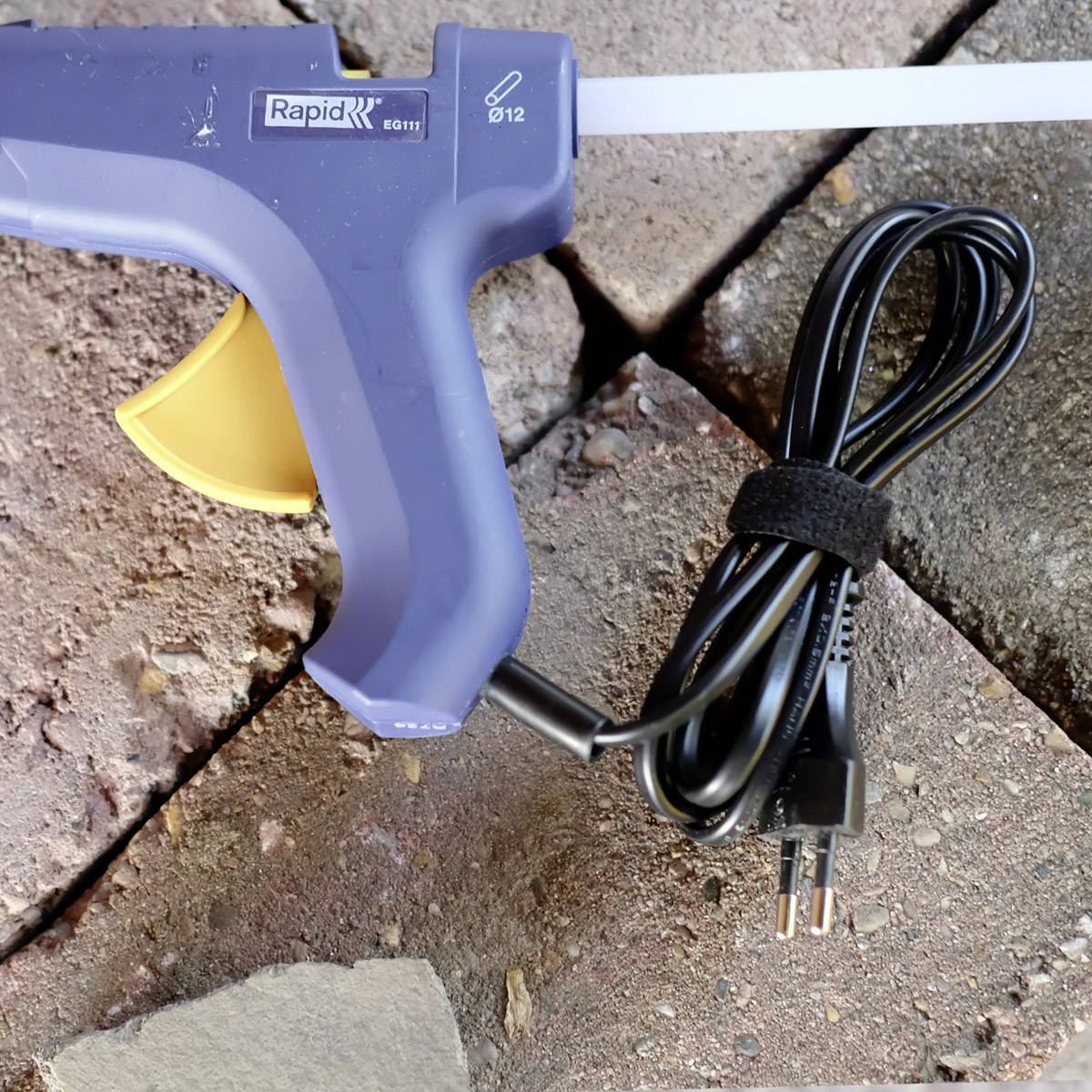 Eine blaue Heißklebepistole mit gelbem Abzug liegt auf einem Ziegelsteinboden. Daneben liegt das Stromkabel ordentlich aufgerollt.